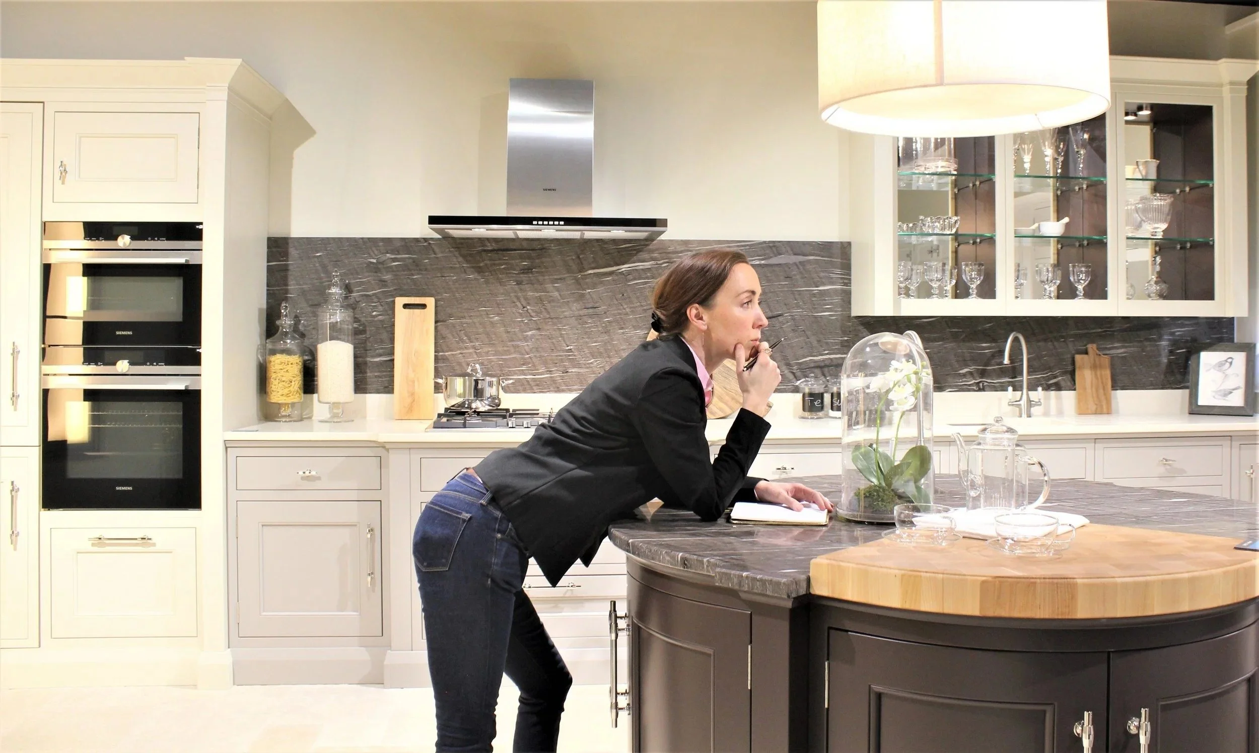 A woman leaning on a kitchen island with her hand on her lips, appearing to be thinking or contemplating, in a modern kitchen setting.