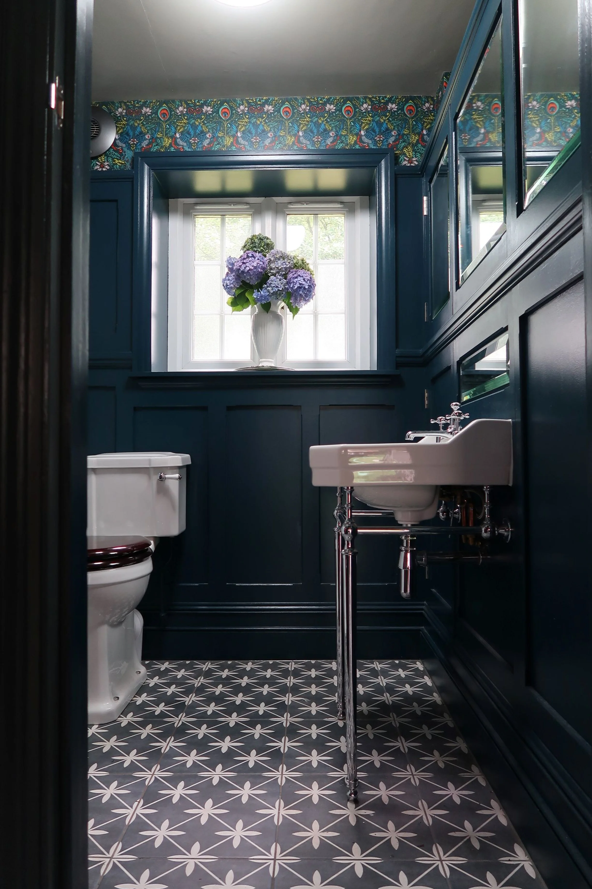 A small bathroom with black paneling, a window with white trim, and a floral bouquet of purple and blue hydrangeas in a white vase on the windowsill. There is a white toilet and a white sink with exposed chrome plumbing. The ceiling is light-colored, and the wall above the paneling has a colorful, intricate peacock feather and floral wallpaper. The floor has black and white patterned tiles.