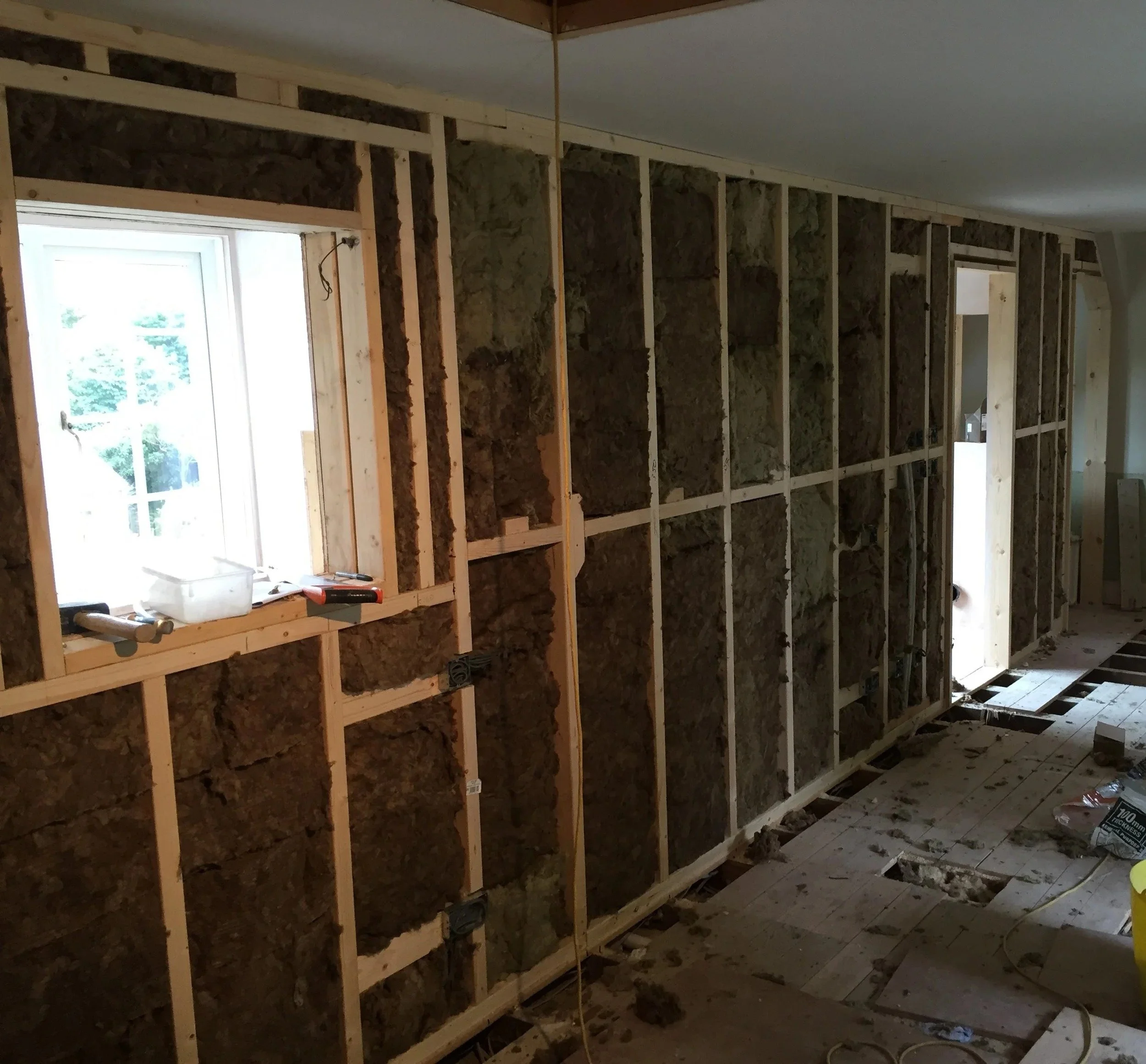Interior view of a room under construction with exposed wooden framing and insulation, window letting in natural light, construction tools and materials on the floor.
