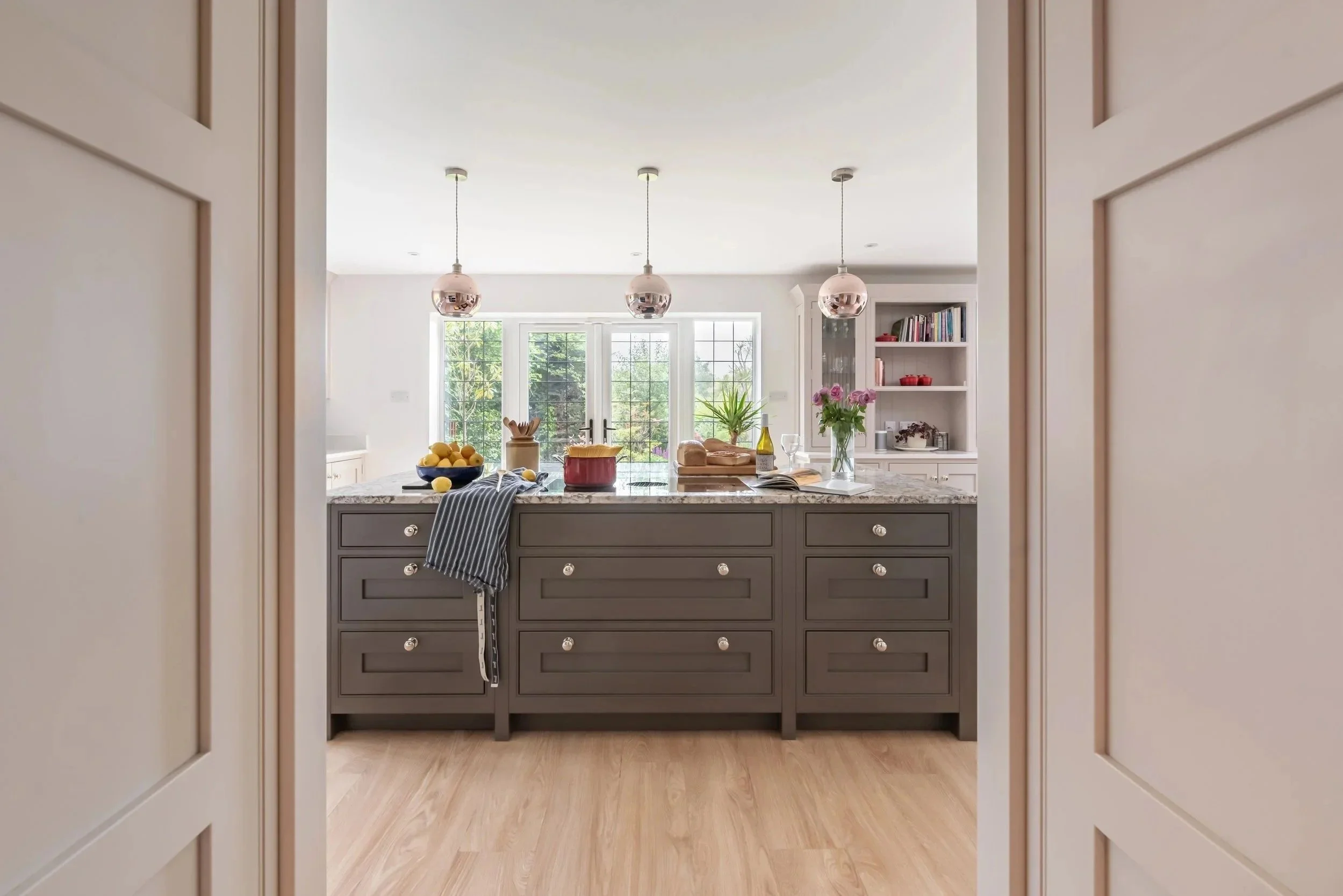 View of a modern kitchen island with gray cabinets, a marble countertop, and various kitchen items including a bowl of lemons, a loaf of bread, a wine bottle, a wine glass, and a flower vase, seen through doorway with white paneled walls, with large windows and a garden outside.