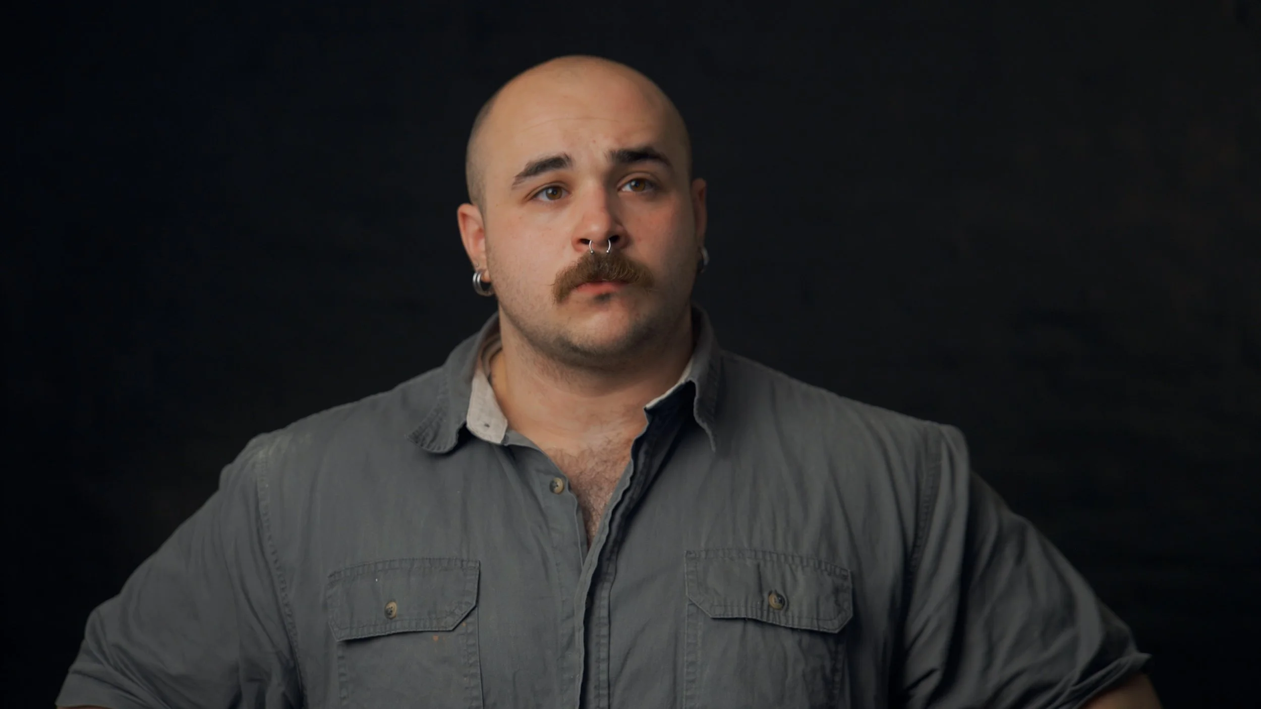 A bald man with a mustache and piercings, wearing a gray button-up shirt, looks slightly to the side against a black background.
