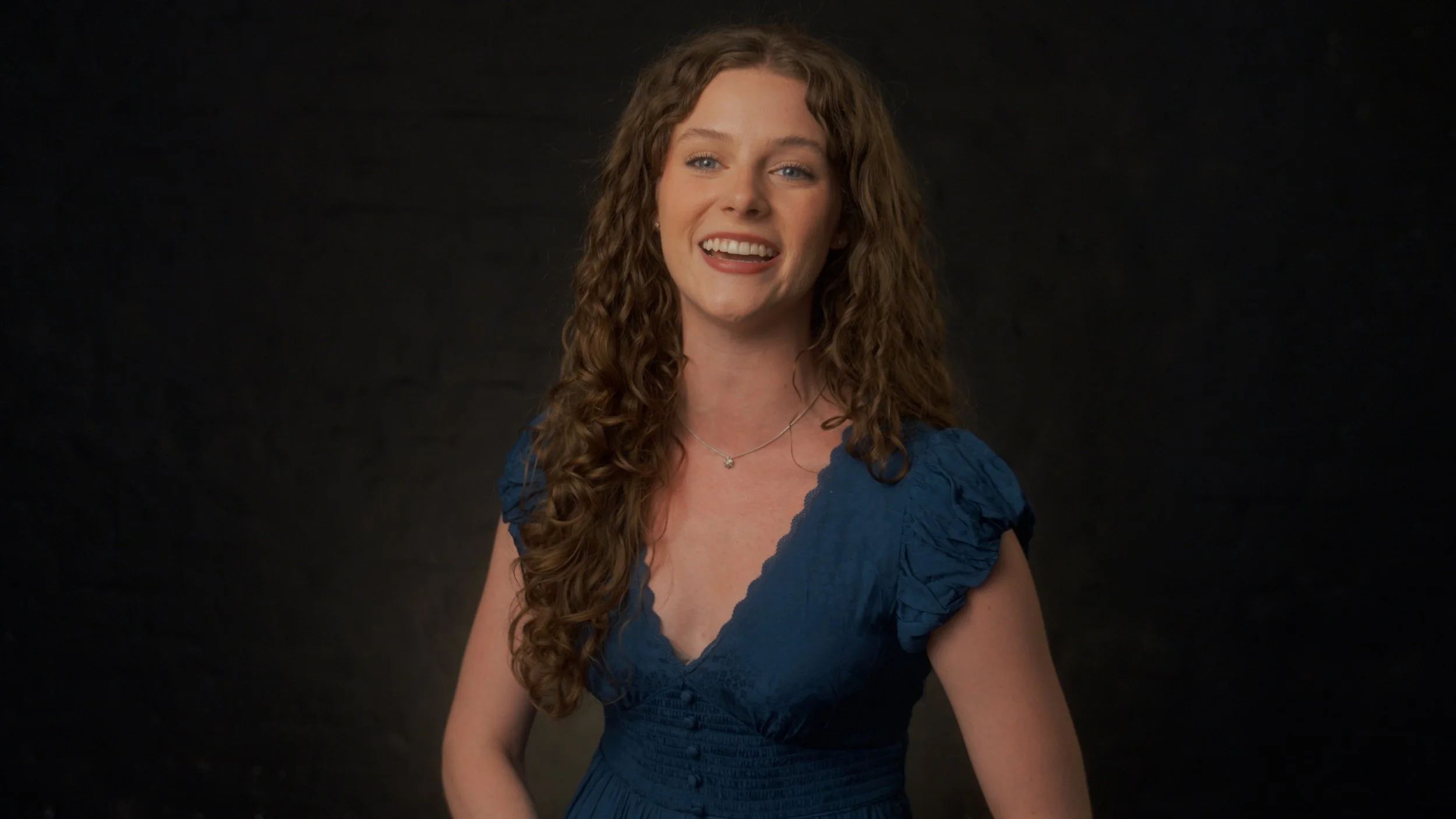 A woman with long, curly auburn hair, wearing a blue dress with puffed sleeves, smiling against a dark background.