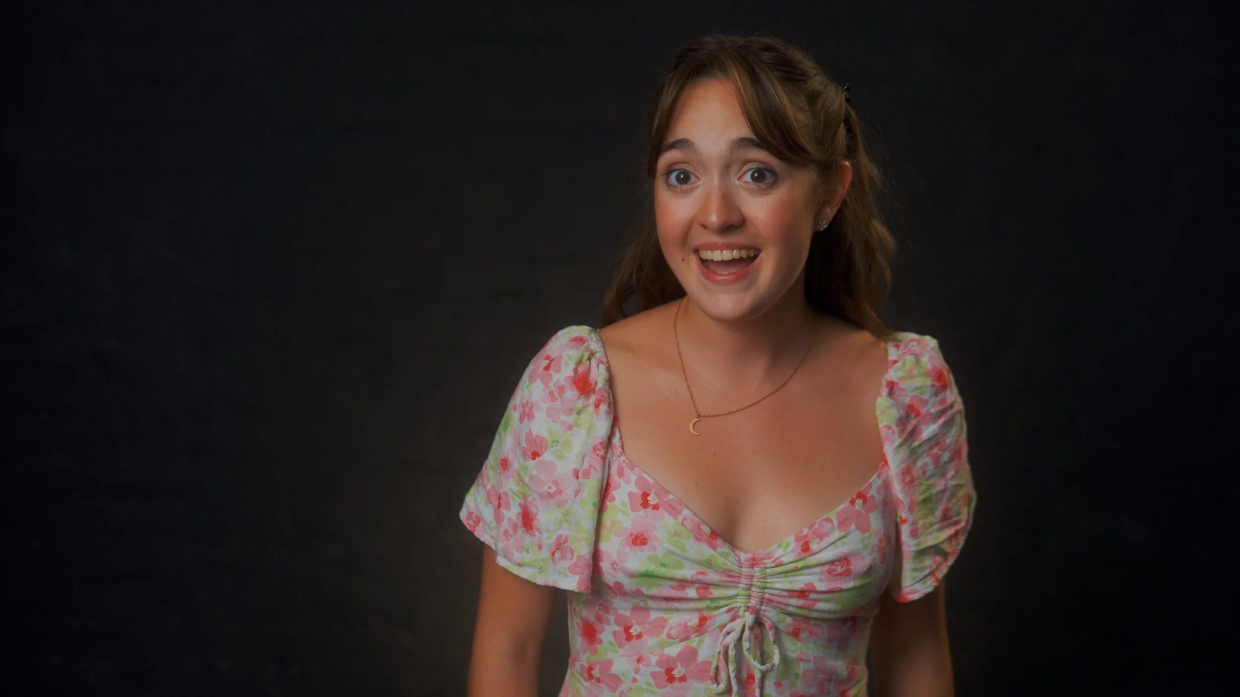 A young woman with brown hair wearing a pink floral dress and a crescent moon necklace, smiling against a dark background.
