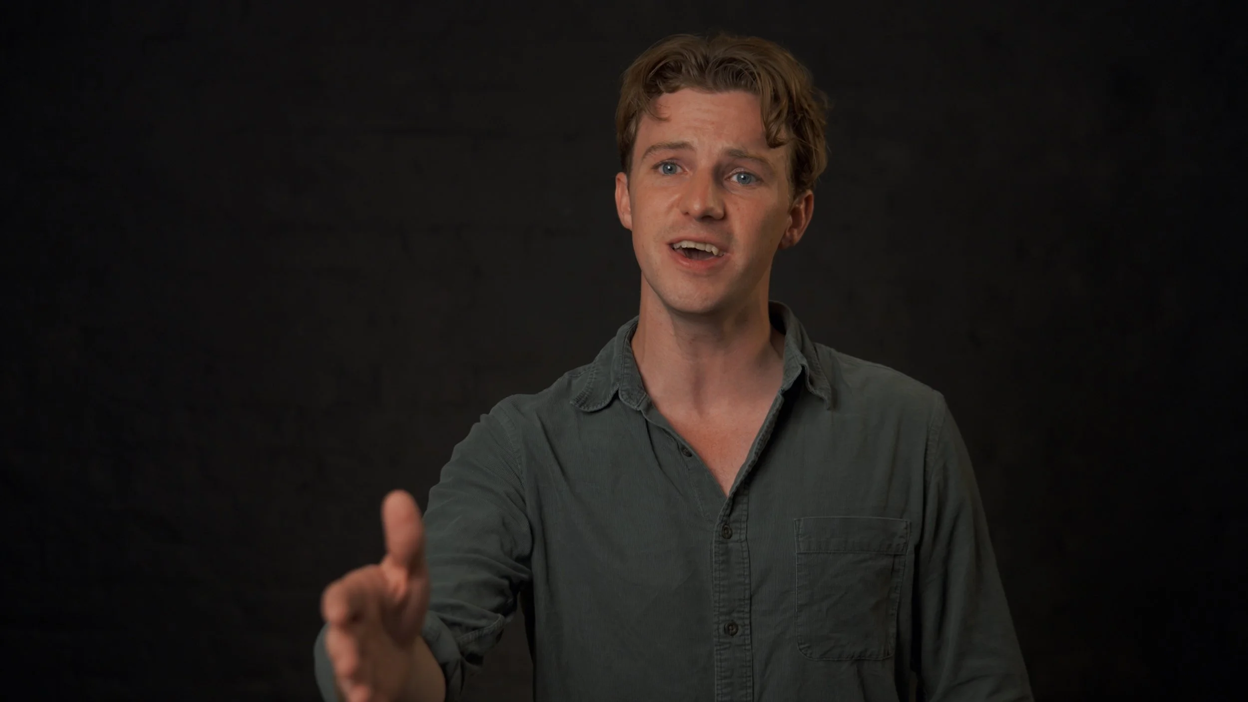 A man with light brown hair wearing a gray button-up shirt, gesturing with his right hand, speaking against a plain dark background.