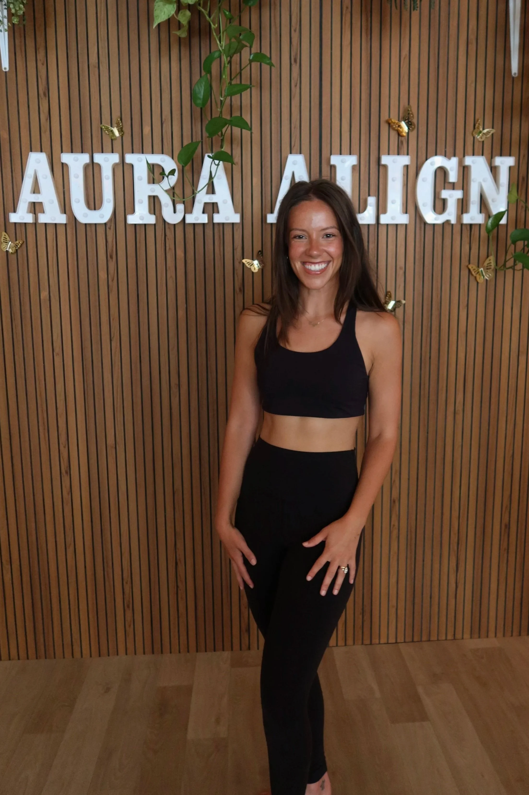 A young woman with long brown hair smiling in a fitness studio, wearing a black sports bra and high-waisted leggings, with neon signage and gym equipment in the background.