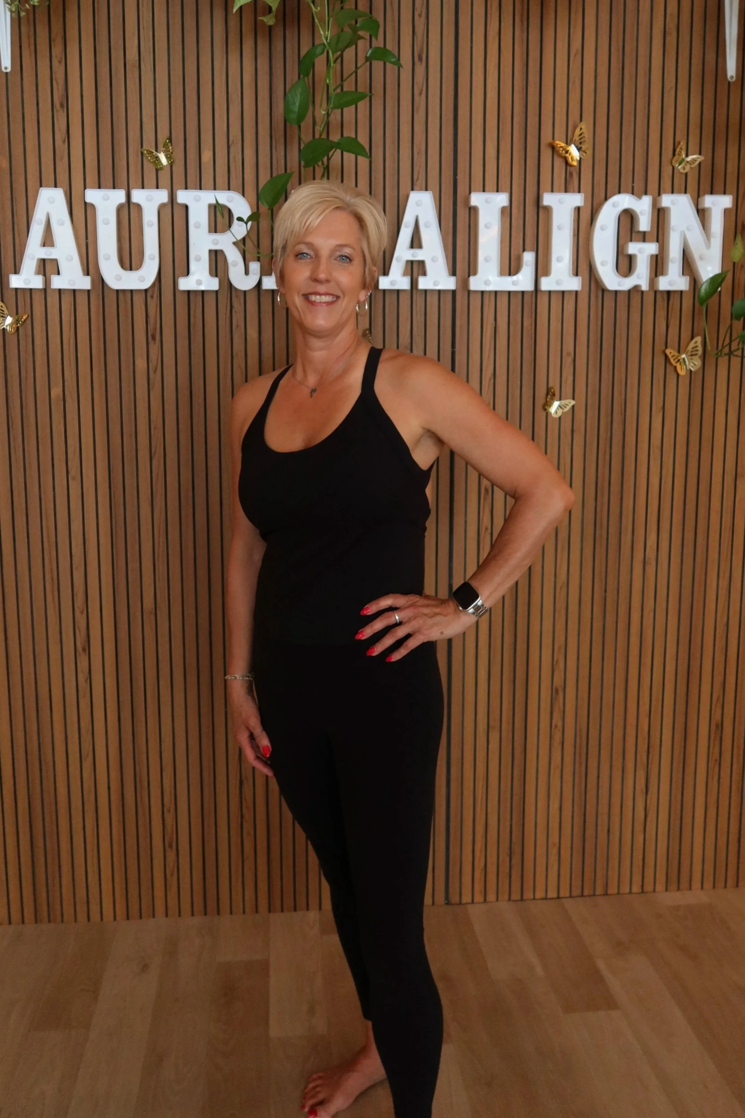 Smiling woman with short blonde hair in a black sports top in a fitness studio, with Pilates equipment and neon sign in the background.
