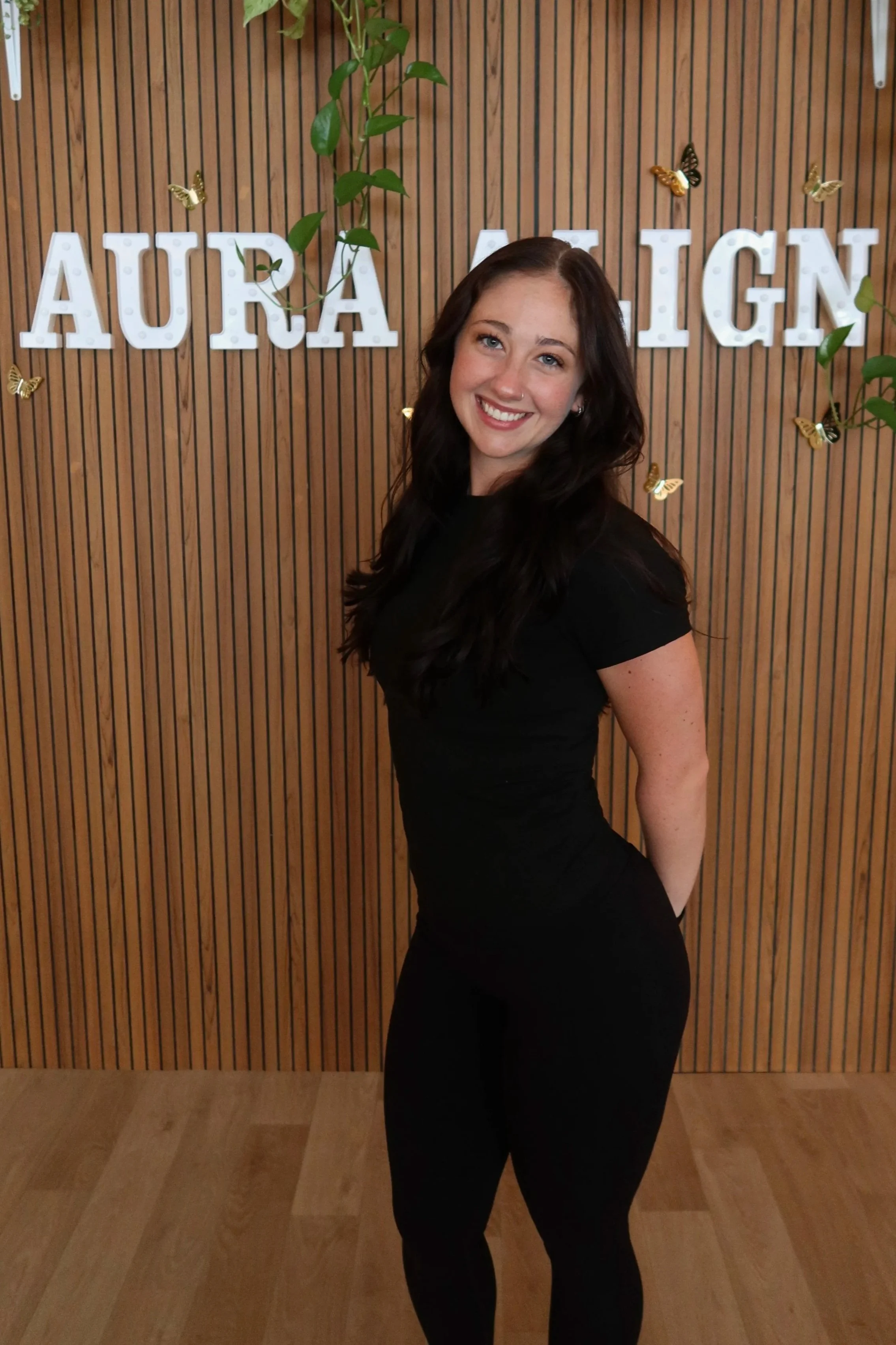 A woman with long dark hair, blue eyes, and a nose ring, smiling in a modern interior space with black walls, neon signs, and industrial lighting.