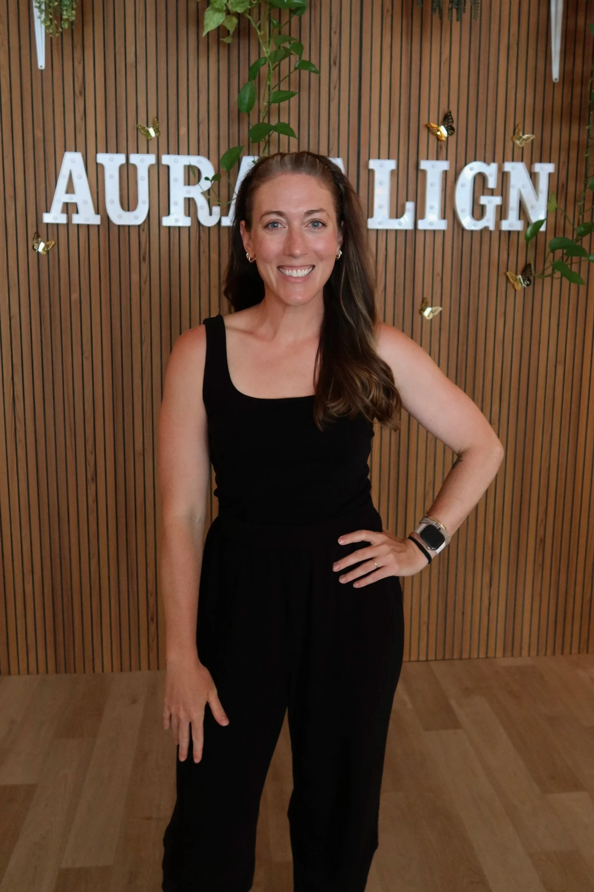 A woman with long wavy brown hair and blue eyes smiling, wearing a black sports top with cutouts, standing in a modern indoor setting with black ceiling lights and illuminated wall decor.