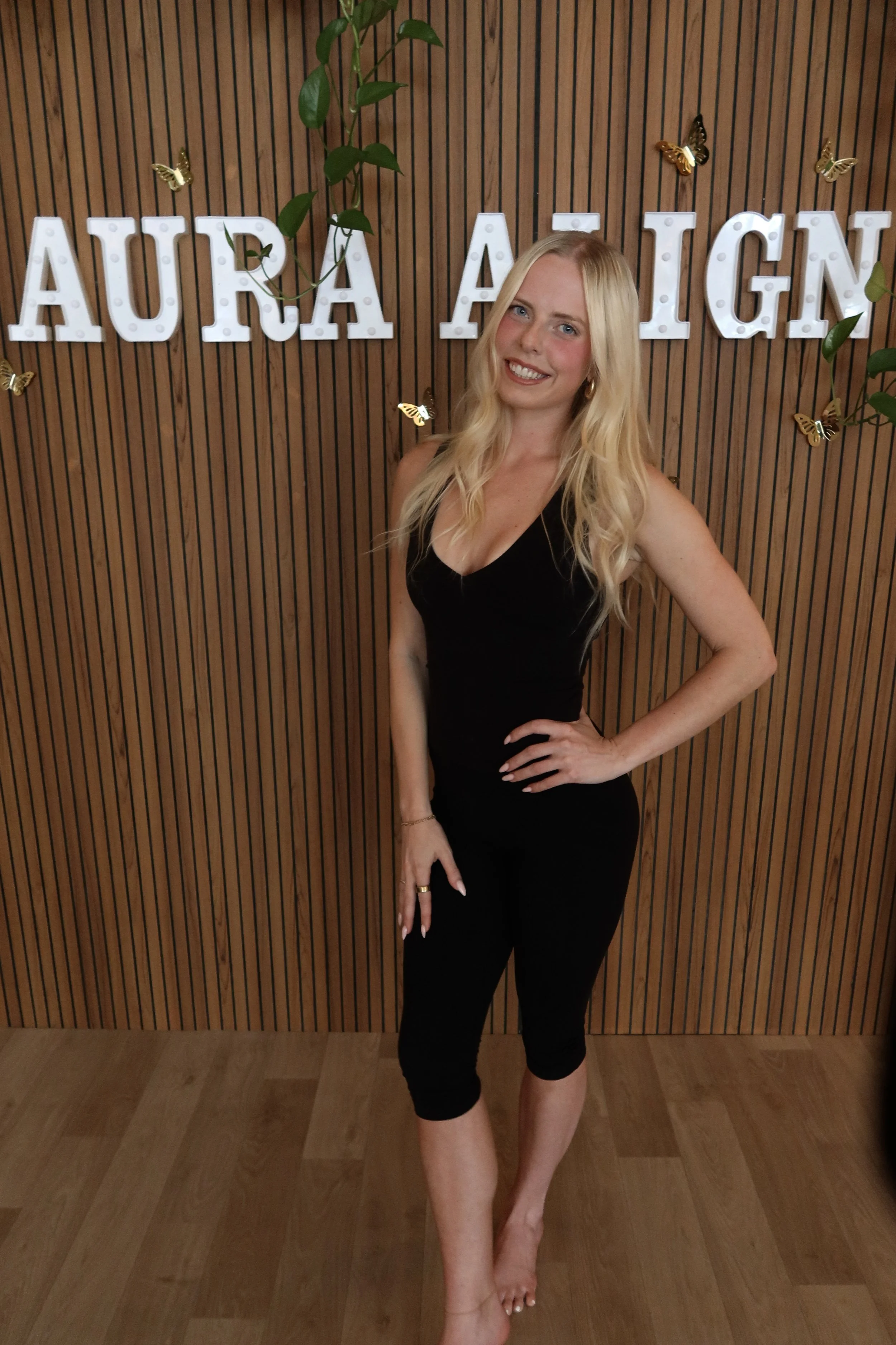 A smiling young woman with long blonde hair wearing a black sleeveless top, gold jewelry, and gold hoop earrings, standing in an indoor space with modern lighting and decor.