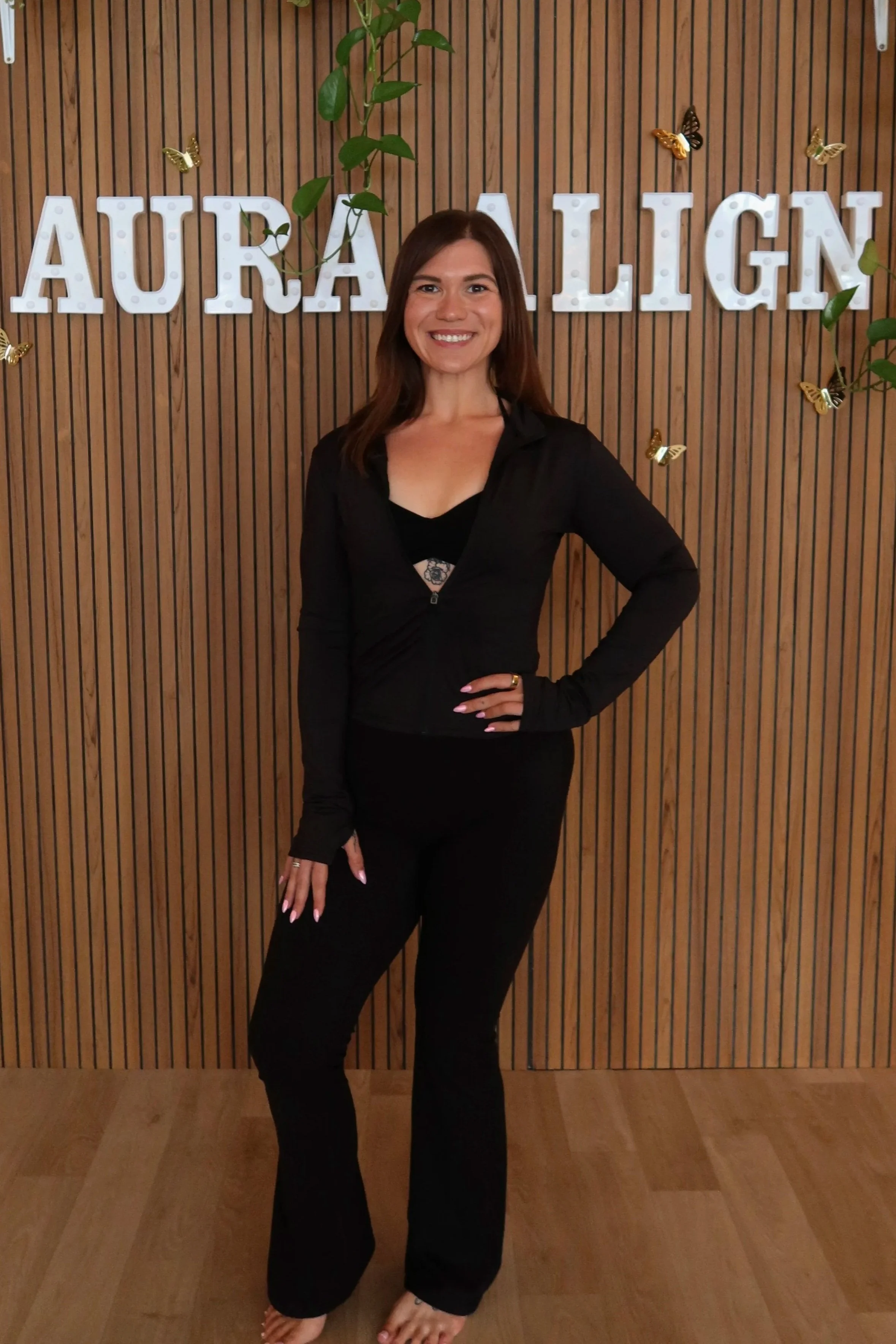 A woman with long, wavy blonde hair, wearing a black, long-sleeved top with a spaghetti strap on her shoulder. She is smiling and standing indoors with modern lighting and decor in the background.