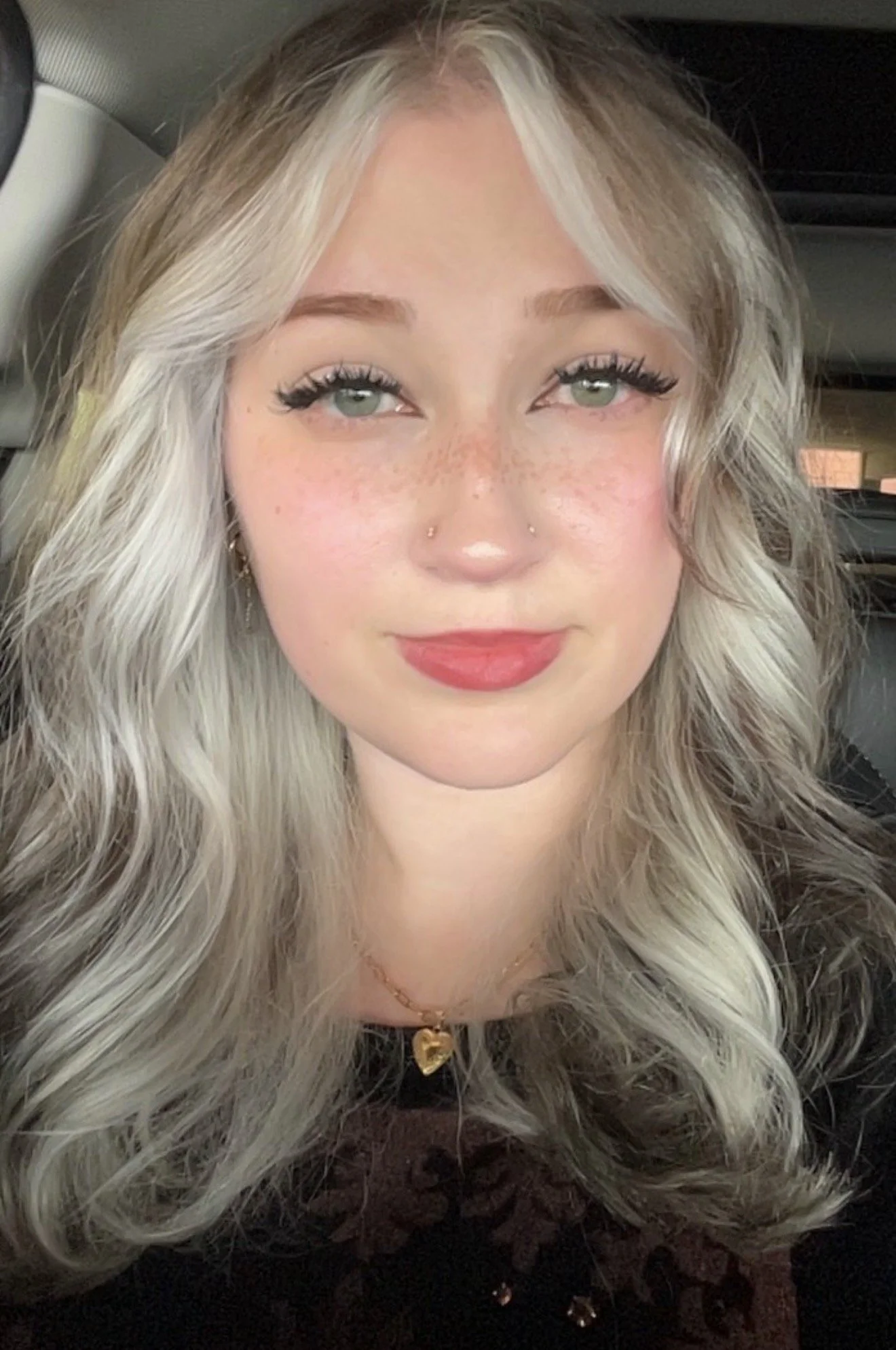 Close-up of a young woman with long wavy platinum blonde hair, green eyes with makeup including mascara and eyeliner, light freckles, wearing a heart-shaped pendant necklace, and a black top.