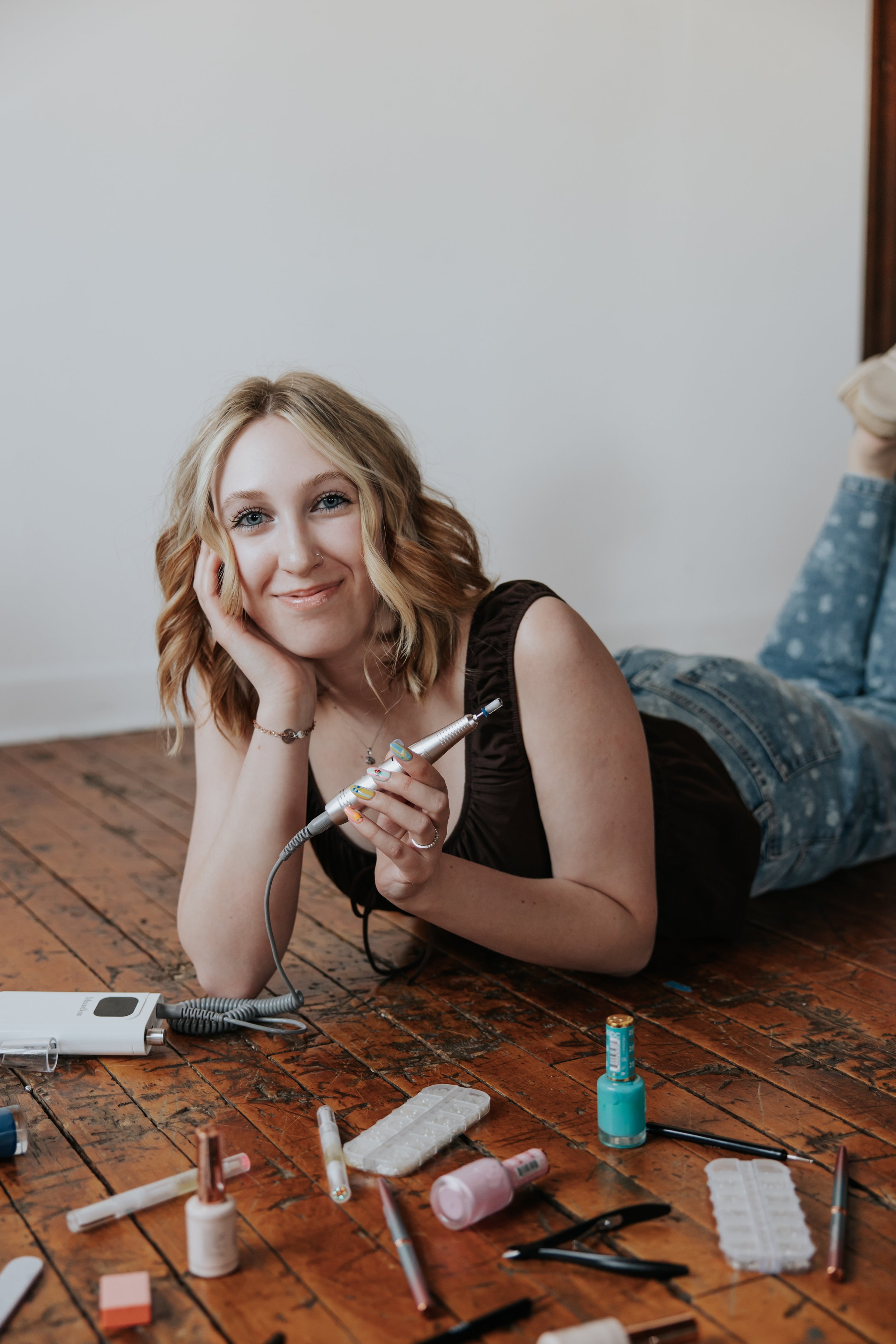 A young woman with blonde hair lying on her stomach on a wooden floor, smiling at the camera while holding a beauty device in her hand. The table in front of her is scattered with various nail polish bottles, tools, and manicure supplies.