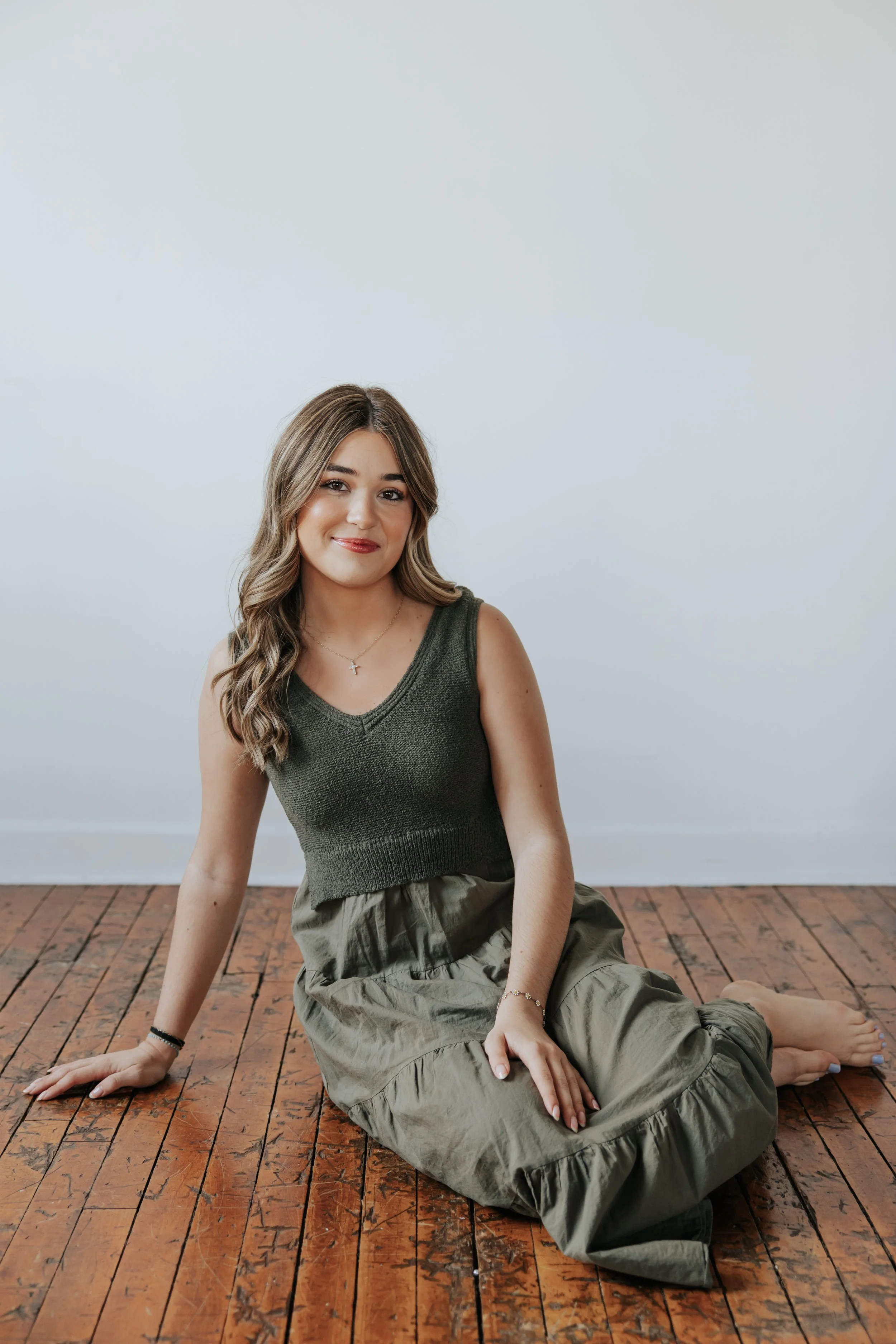 A young woman with styled hair, wearing a sleeveless green top and khaki pants, sitting on a polished wooden floor against a plain light gray wall, smiling at the camera.
