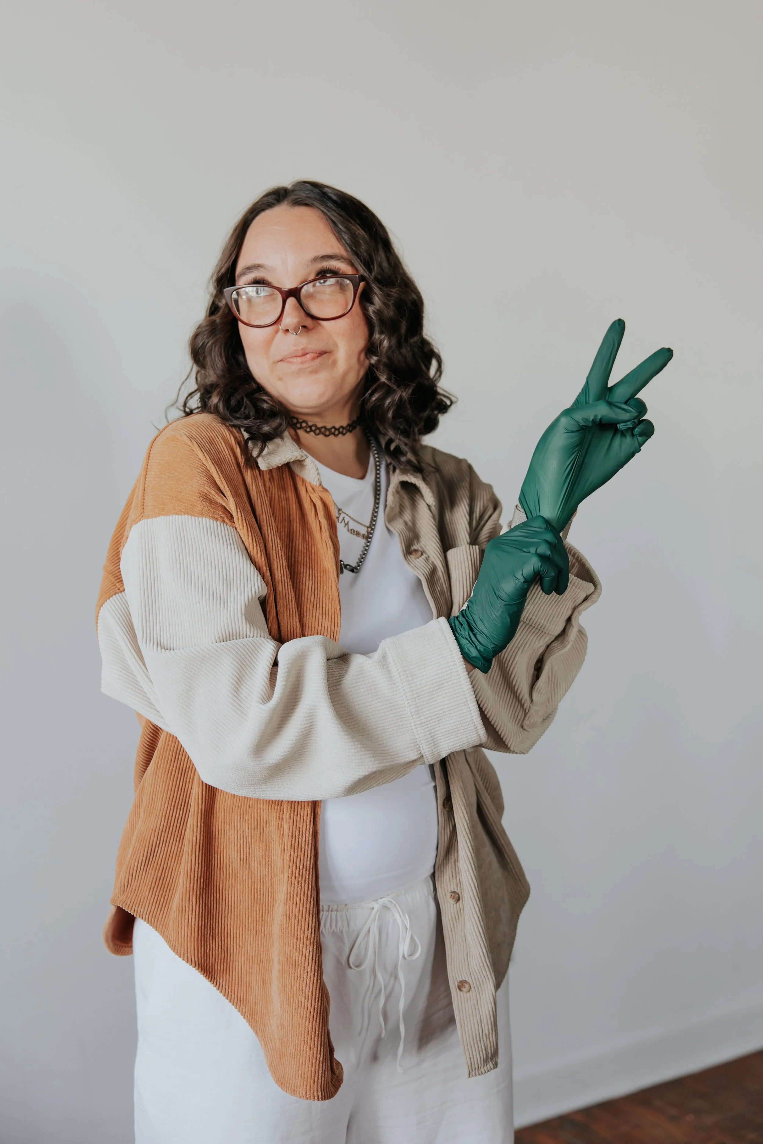 A woman with dark curly hair, glasses, and a septum piercing poses with a peace sign gesture. She is wearing a black choker, layered necklaces, a white t-shirt, a tan and beige corduroy jacket, green gloves, and white pants. She is standing against a plain, light-colored wall.