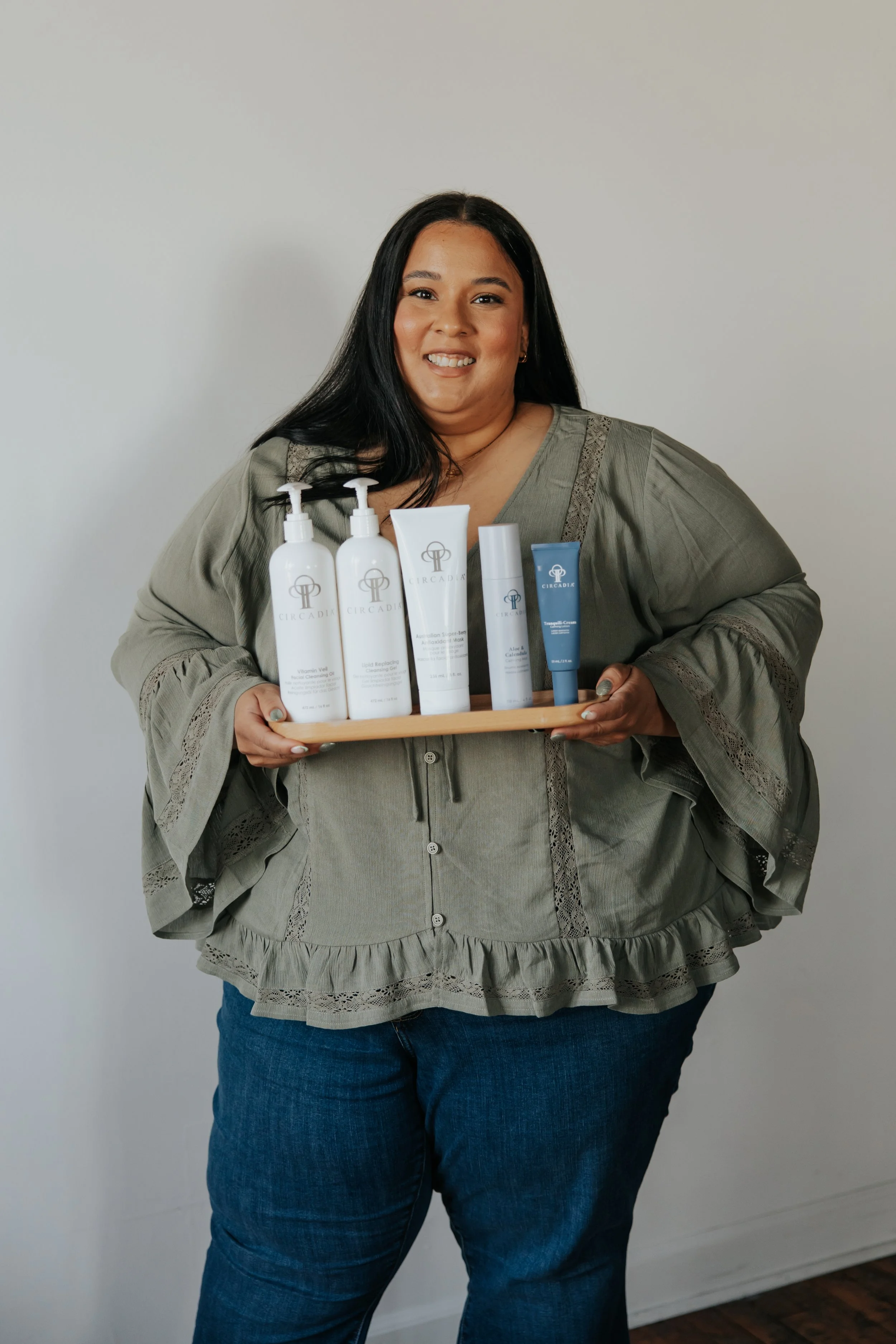 A woman with long black hair and a tan complexion holding a tray with skincare products, wearing a gray blouse with ruffled details and dark blue jeans, standing in front of a plain gray wall.