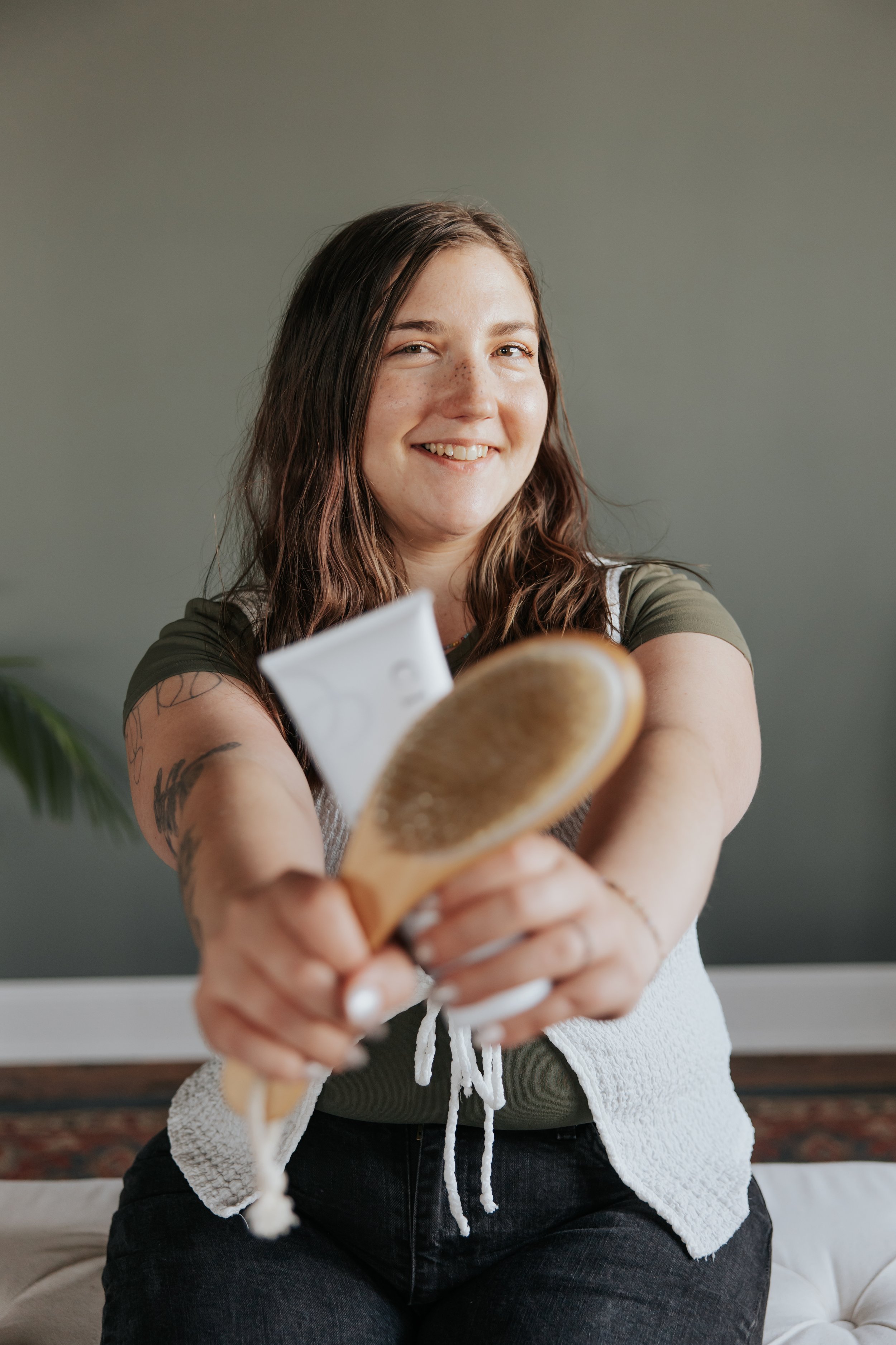 A woman with long brown hair, wearing a green shirt and a white textured vest, smiles while holding a wooden brush towards the camera. The background is a plain wall, and she has visible tattoos on her left arm.