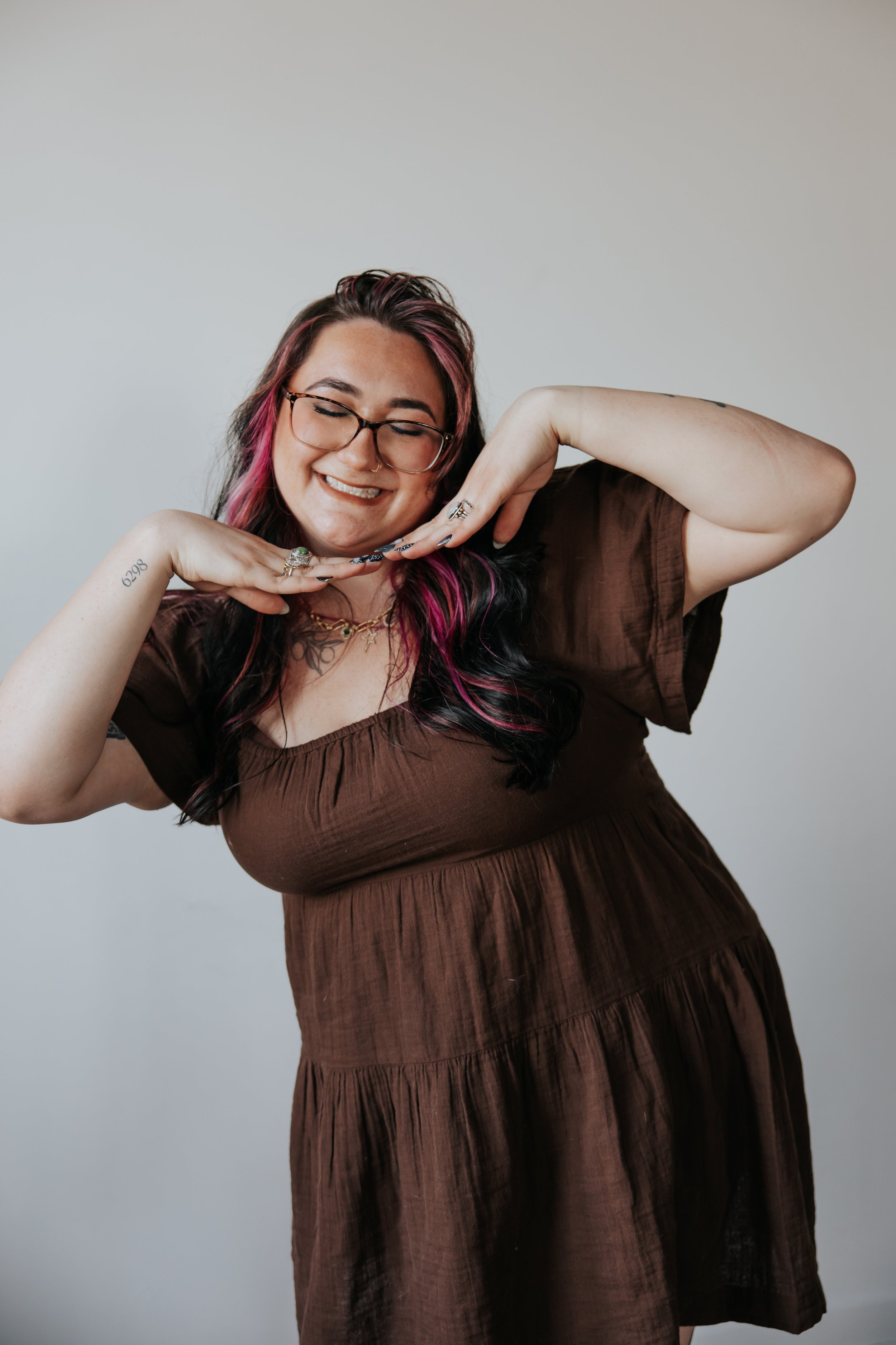 A smiling woman with glasses, pink and black hair, wearing a brown dress, posing with her hands under her chin against a plain white background.