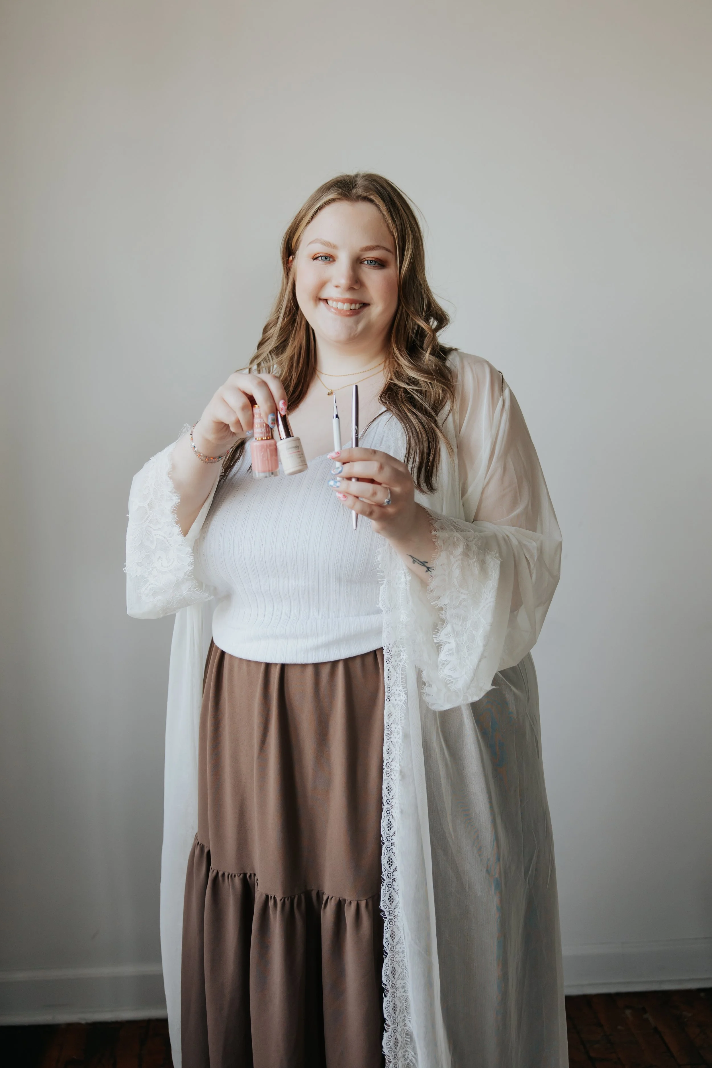 A smiling woman with long wavy brown hair wearing a white top, brown tiered skirt, and a sheer white lace robe, holding nail polish bottles and a nail file in front of a plain light gray wall.