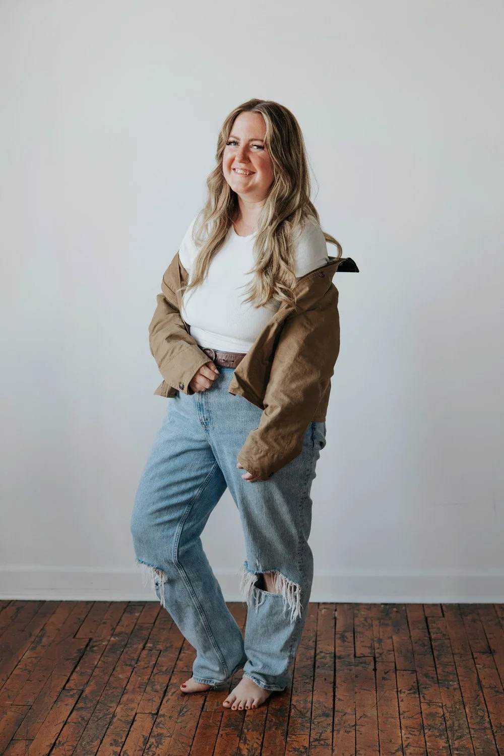 A woman with long wavy hair smiling, standing barefoot on a wooden floor against a plain white wall, dressed in a white T-shirt, a beige jacket, and ripped light blue jeans.