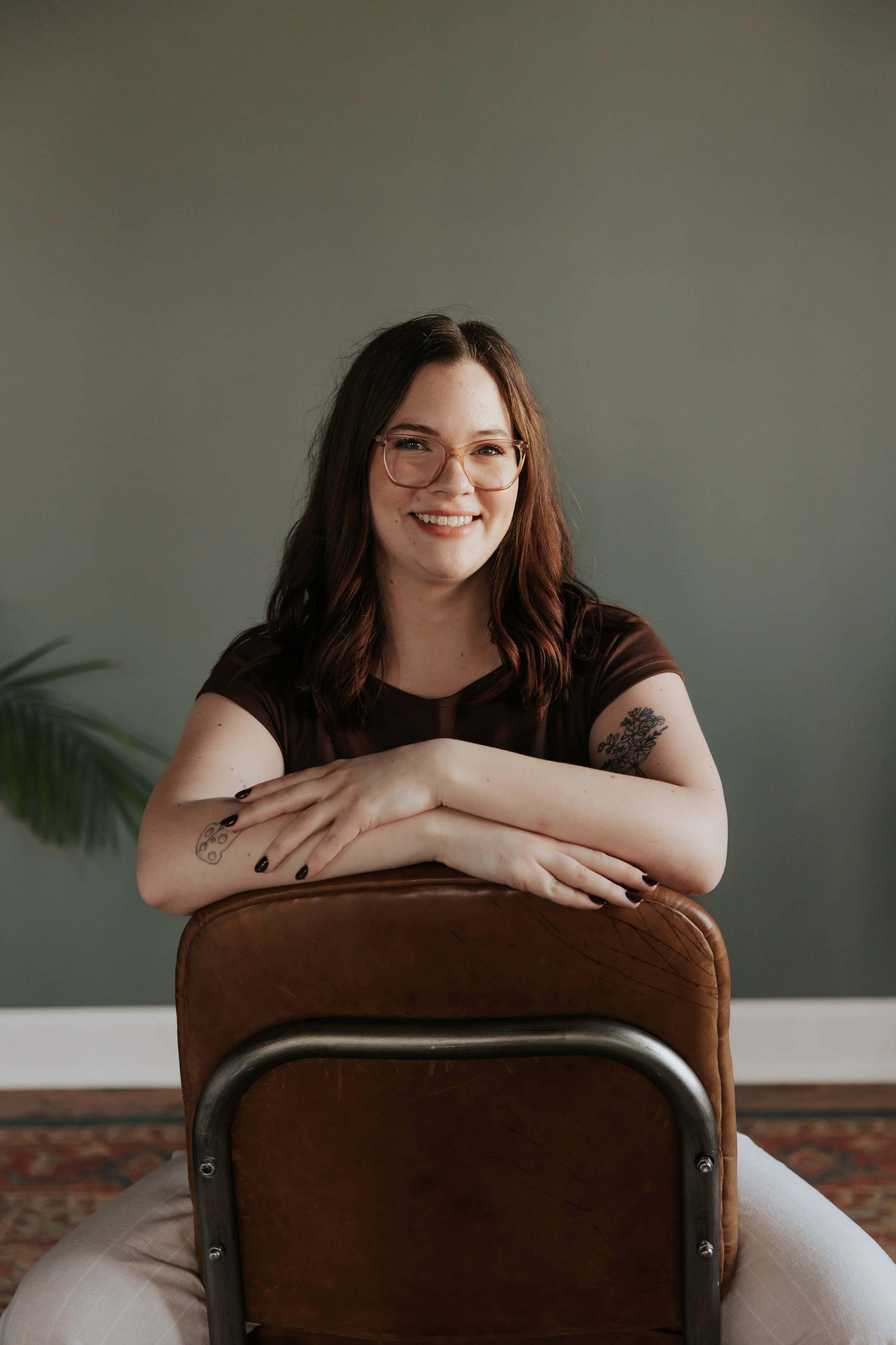 A young woman with brown hair and glasses smiling, sitting on a chair with her arms resting on the back of another chair, in a room with a gray wall and a plant partly visible on the left.