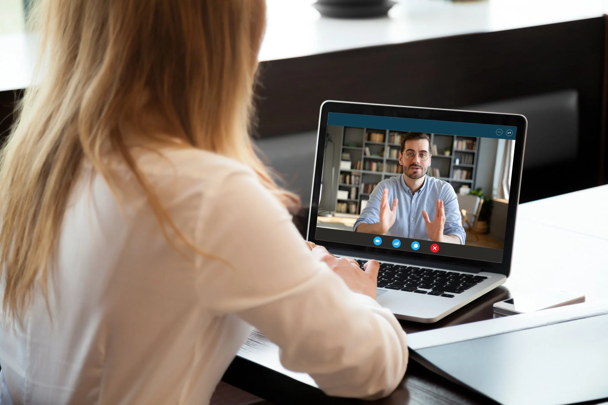A woman with red hair in a white shirt participating in a video call on her laptop with a man in a blue shirt who appears to be explaining something from a home or office setting.