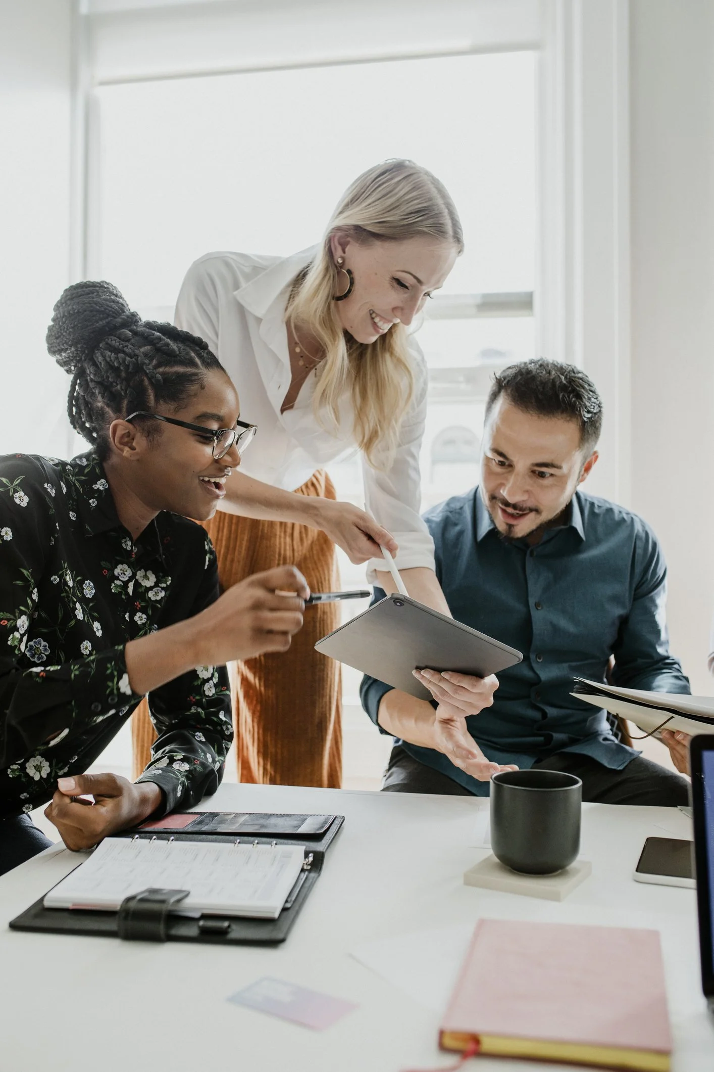 Three coworkers collaborating in a bright office, looking at a tablet and discussing, with notebooks and a coffee mug on the table.