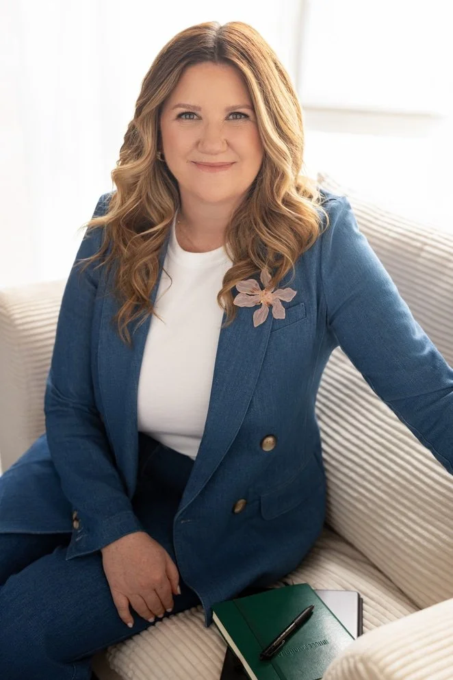 A woman with wavy, shoulder-length blonde hair, wearing a blue blazer with a pink flower pin, sitting on a beige sofa with a green notebook and black pen, smiling at the camera in a well-lit room.