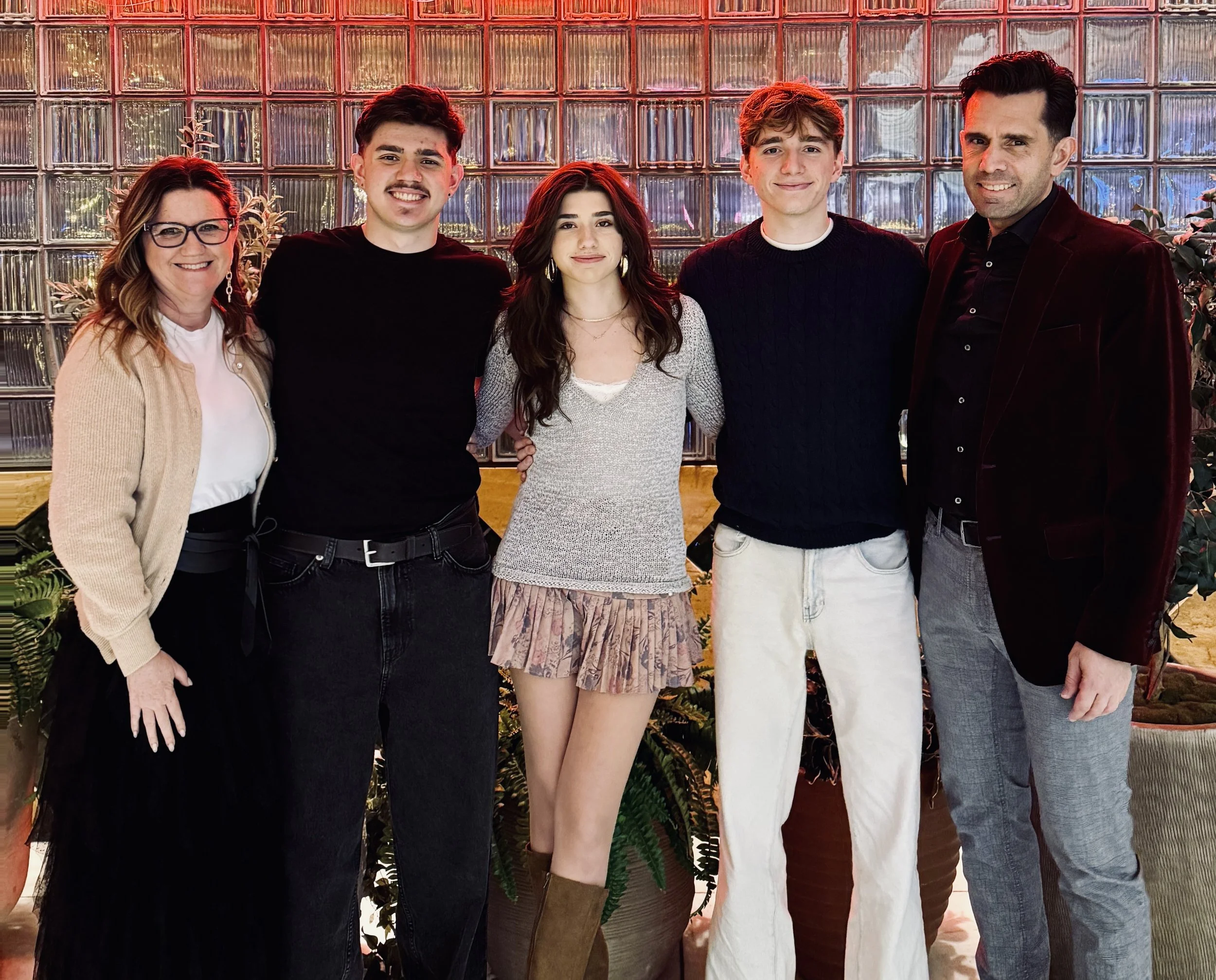 Lauire J Wetzel and her family standing together in front of a glass block wall, smiling at the camera. They are indoors with potted plants around them.