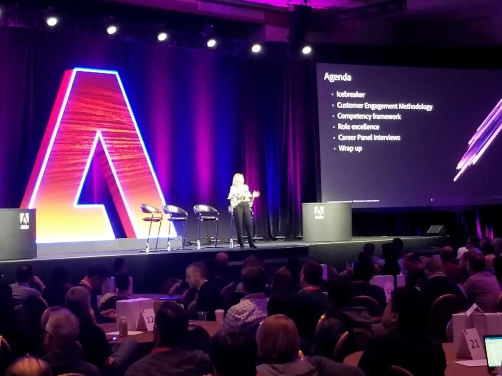 A woman presenting on stage at a conference with a large abstract neon-like 'A' display and a screen displaying an agenda including topics like icebreaker, customer engagement methodology, competency framework, role excellence, career panel interviews, and wrap up. Audience members seated at tables are watching the presentation.