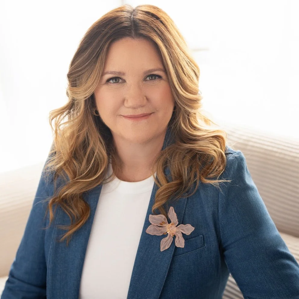 Laurie J Wetzel with long wavy auburn hair, wearing a denim blazer with a pink flower pin, smiling at the camera in a well-lit room.