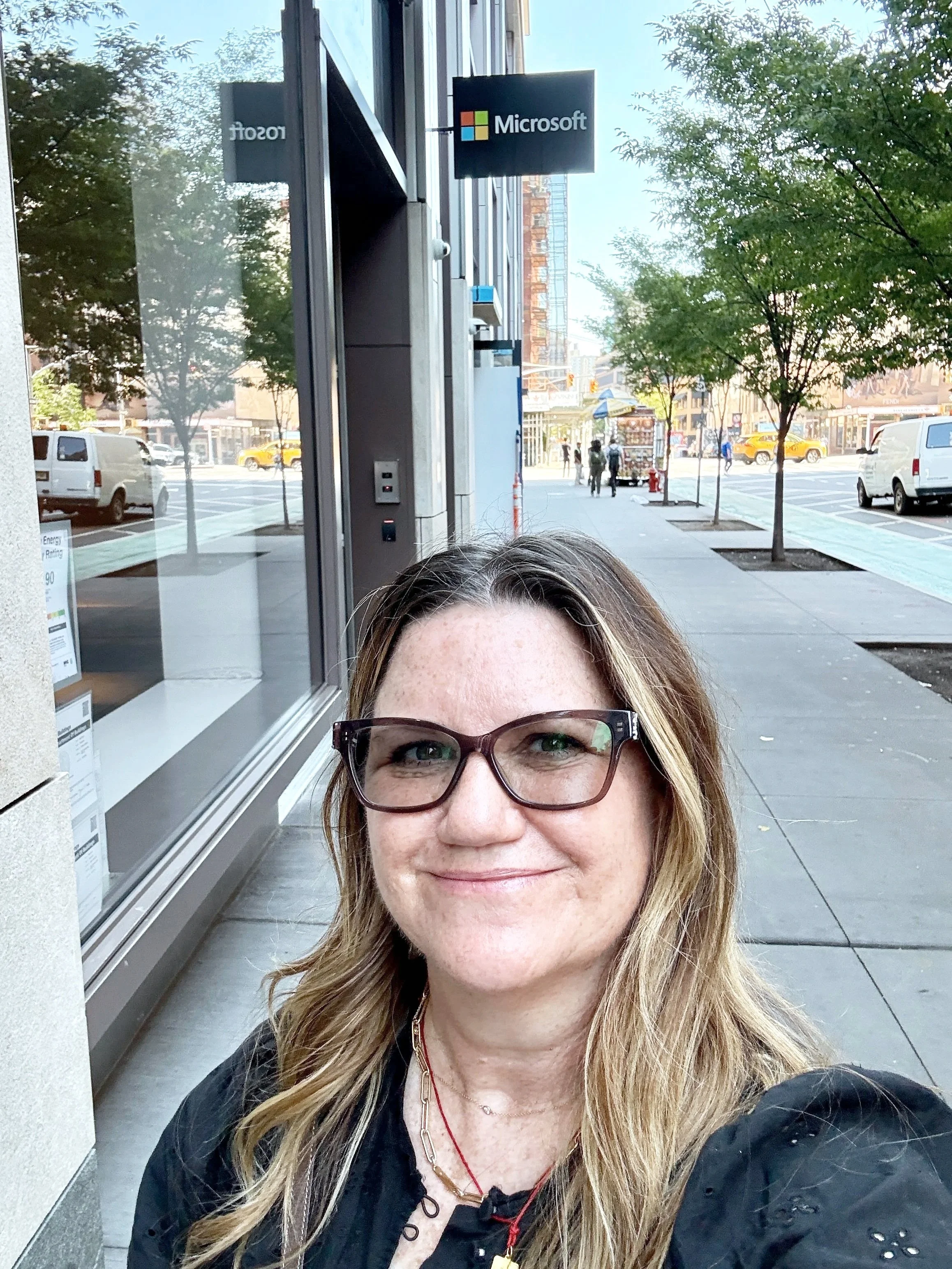 A woman with glasses taking a selfie in front of a Microsoft store on a city sidewalk. The street has trees, parked cars, and yellow taxis in the background.
