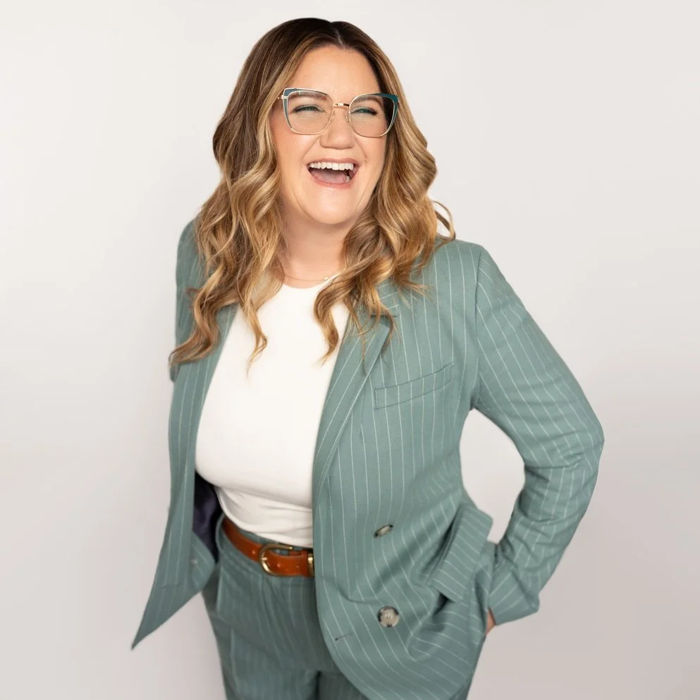 A woman with wavy, shoulder-length hair, wearing glasses, a green pinstripe suit, and a white top, laughing with her eyes closed, against a plain white background.