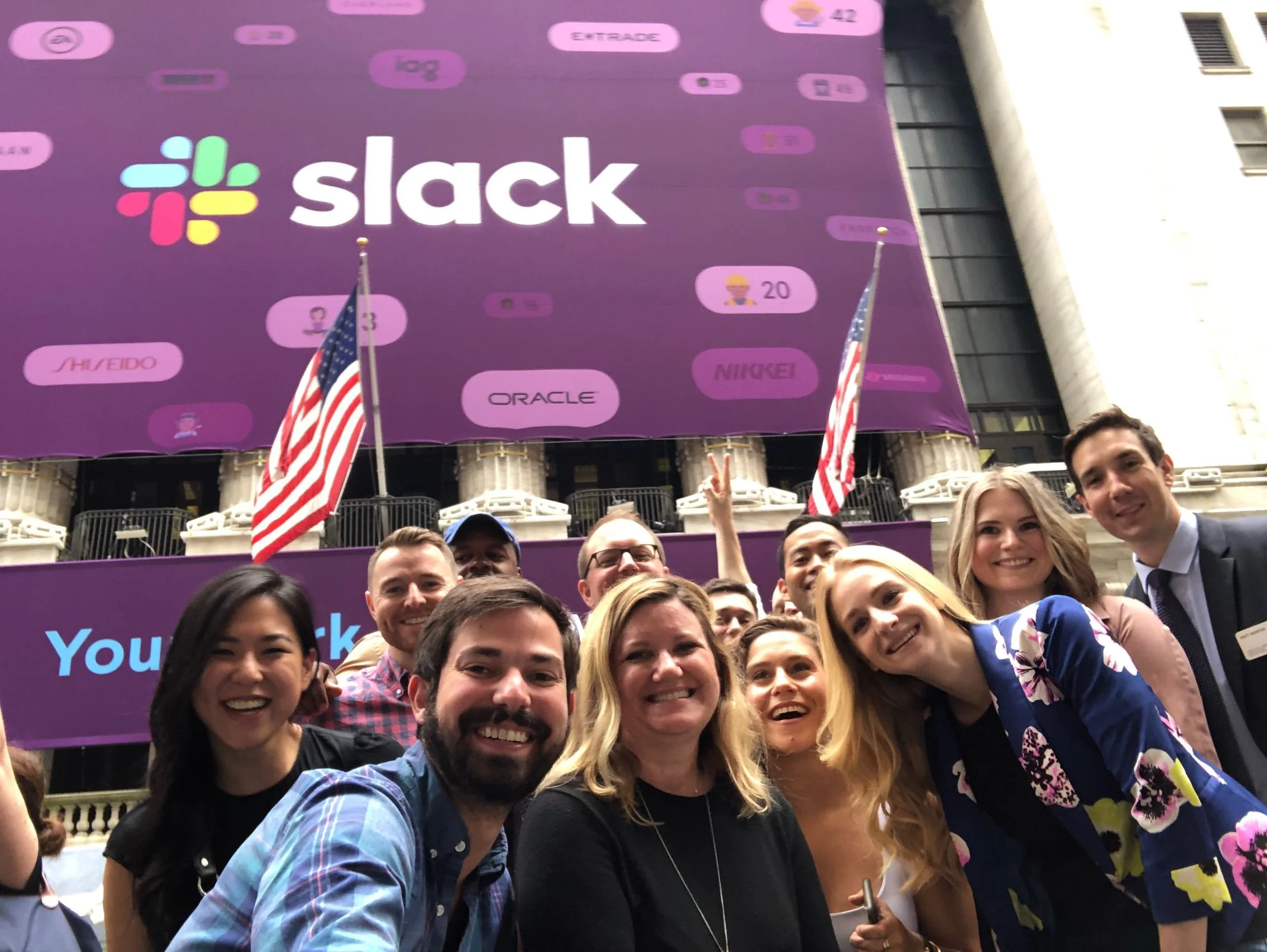 Group of people taking a selfie in front of a large screen displaying Slack logo and logos of various companies at a financial district, with American flags.
