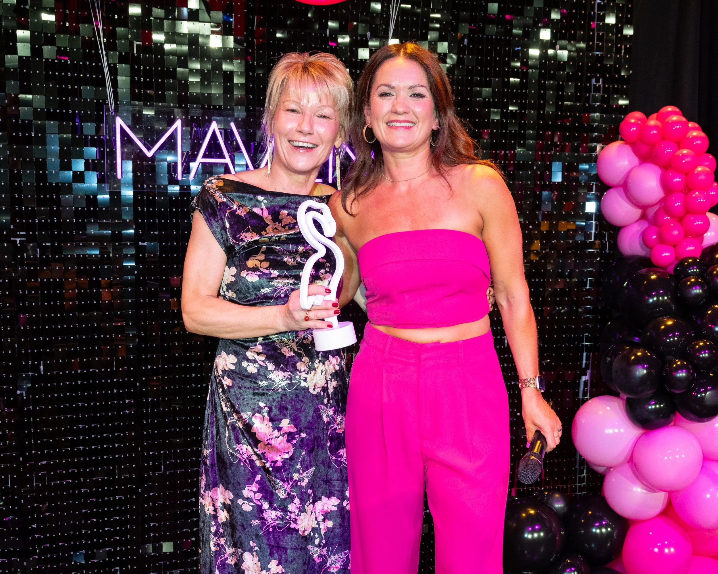 Two women smiling, one holding a flamingo-shaped neon light, standing in front of a colorful, reflective backdrop with pink and black balloons on the right side.