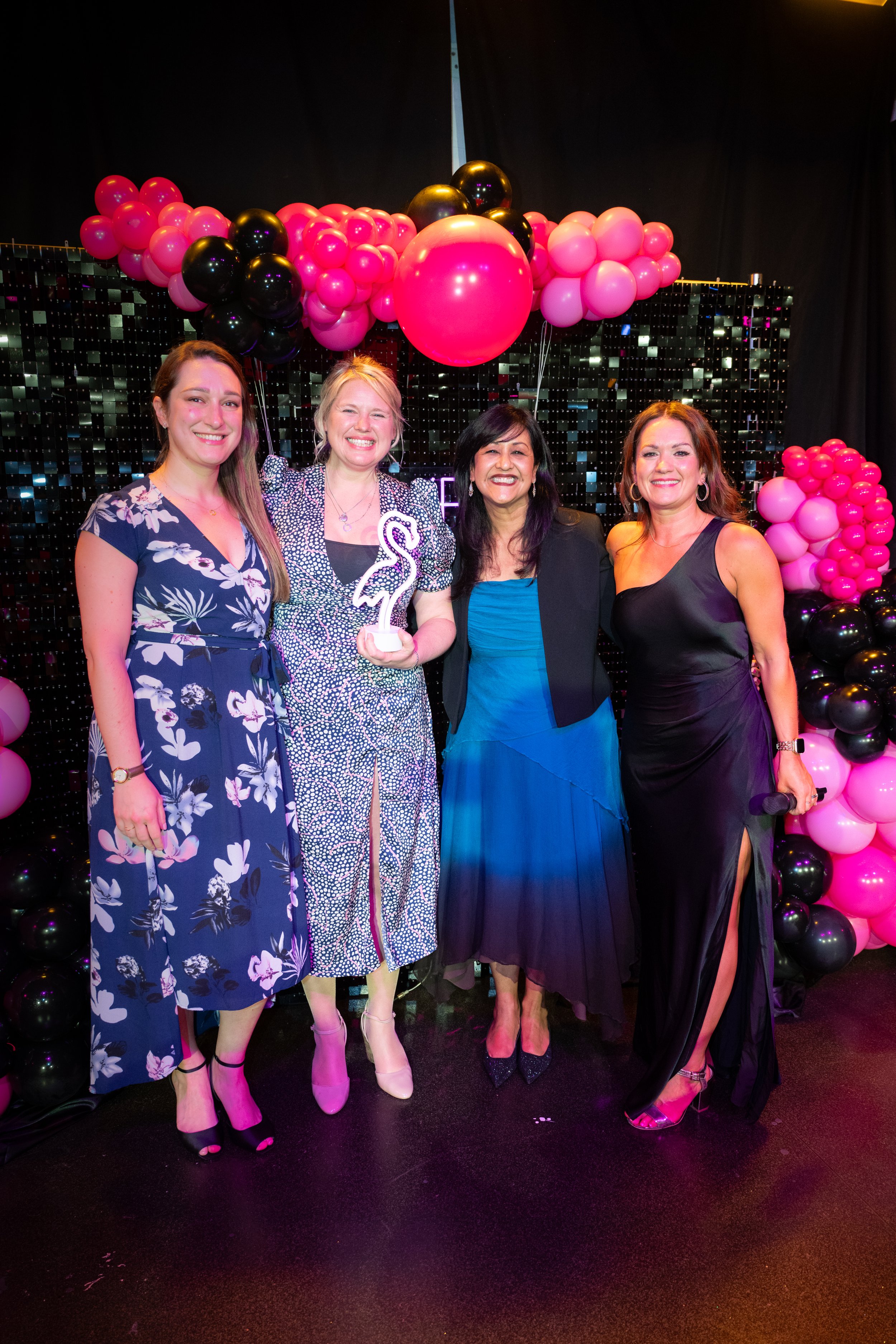 Four women in dresses posing together, smiling, at an event with pink and black balloon decorations behind them.