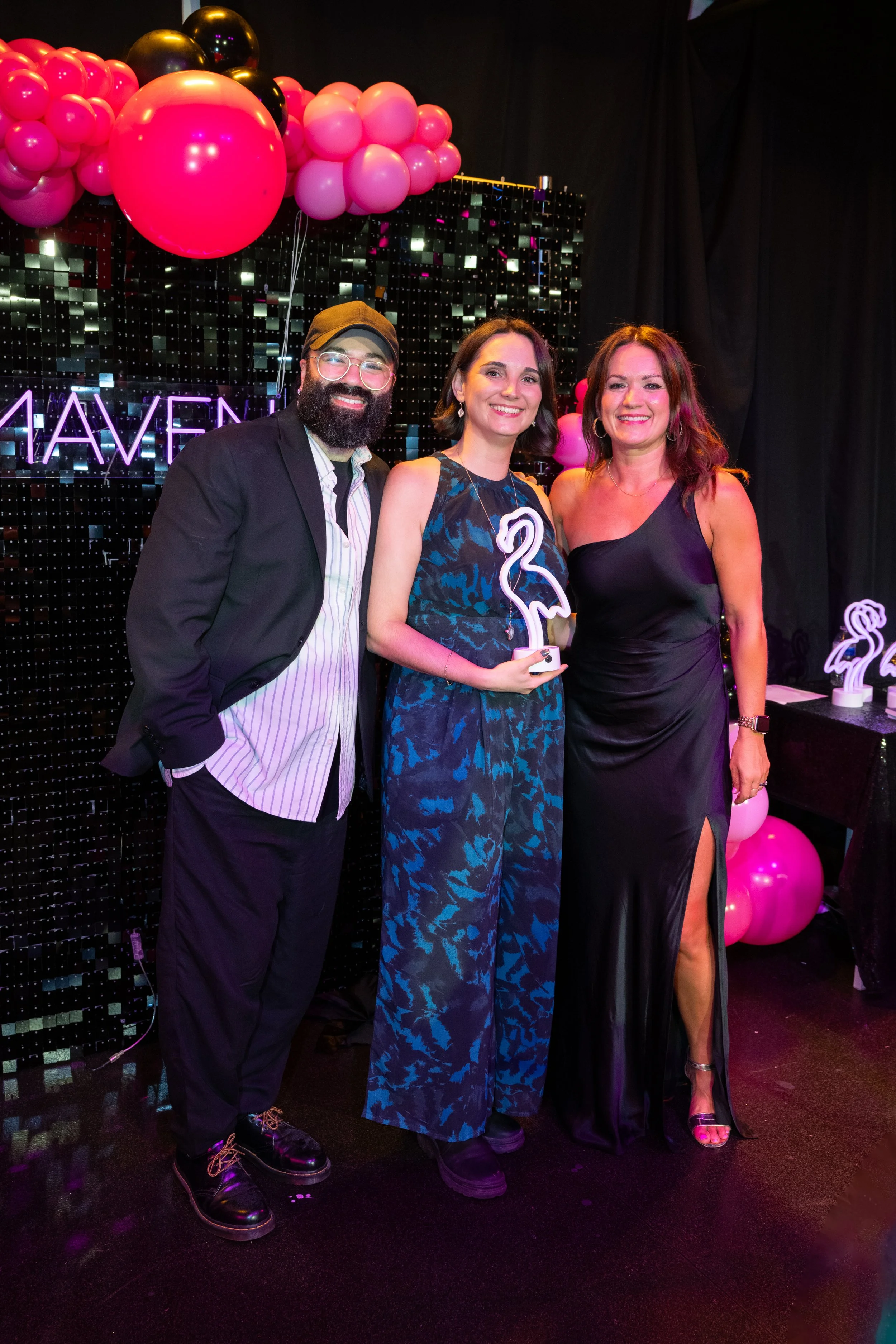 Three people standing together at a celebration event with pink and black balloons, pink neon sign, and a sparkly black backdrop in the background. The man on the left is wearing glasses, a black blazer, and a striped shirt. The woman in the middle is holding a neon flamingo decoration and wearing a blue patterned dress. The woman on the right is in a black dress with a high slit, wearing hoop earrings and a watch.