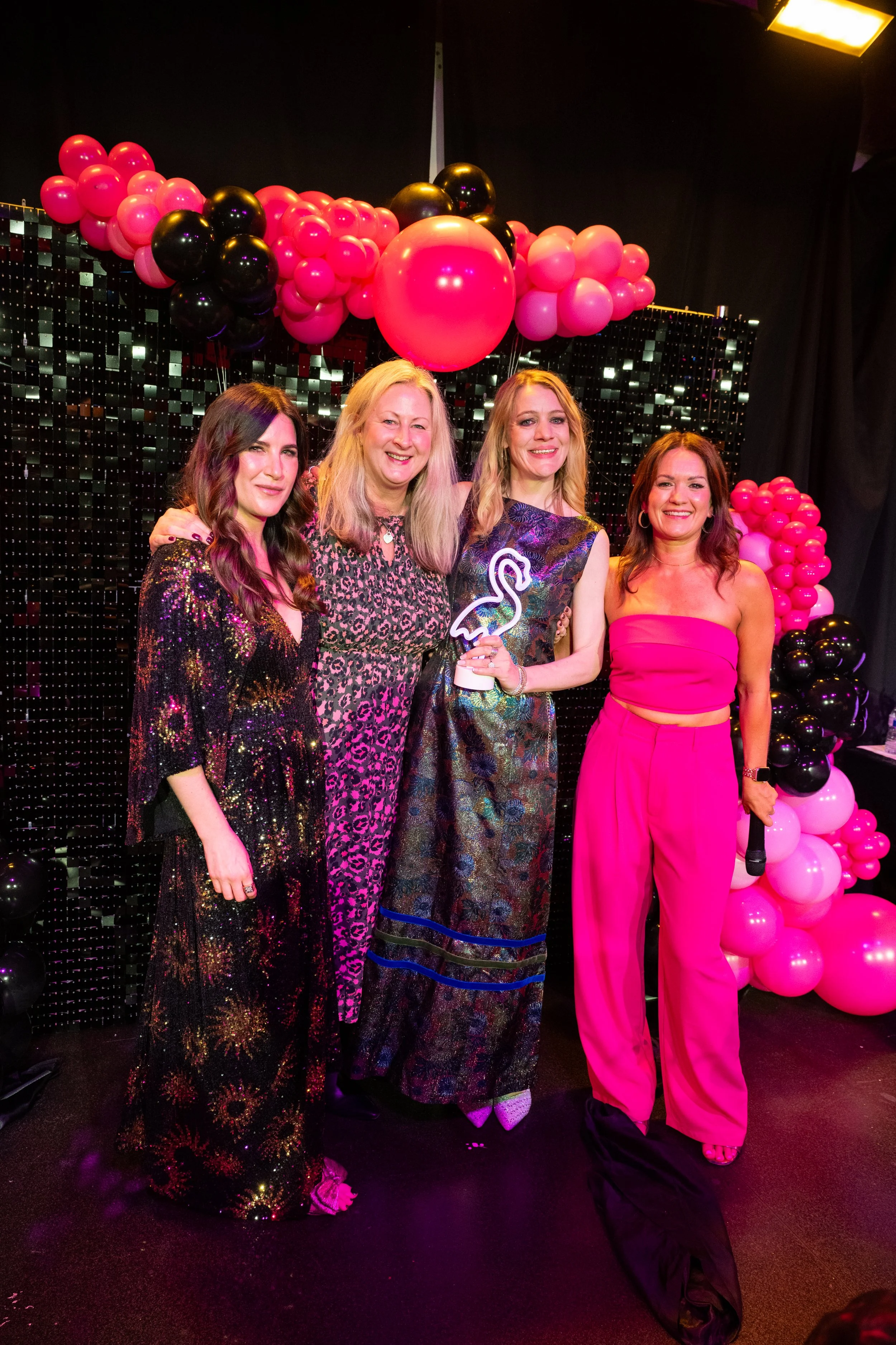 Four women dressed in colorful, party attire standing together at a celebration event, with a backdrop of pink, black, and white balloon decorations and a sparkly black curtain.