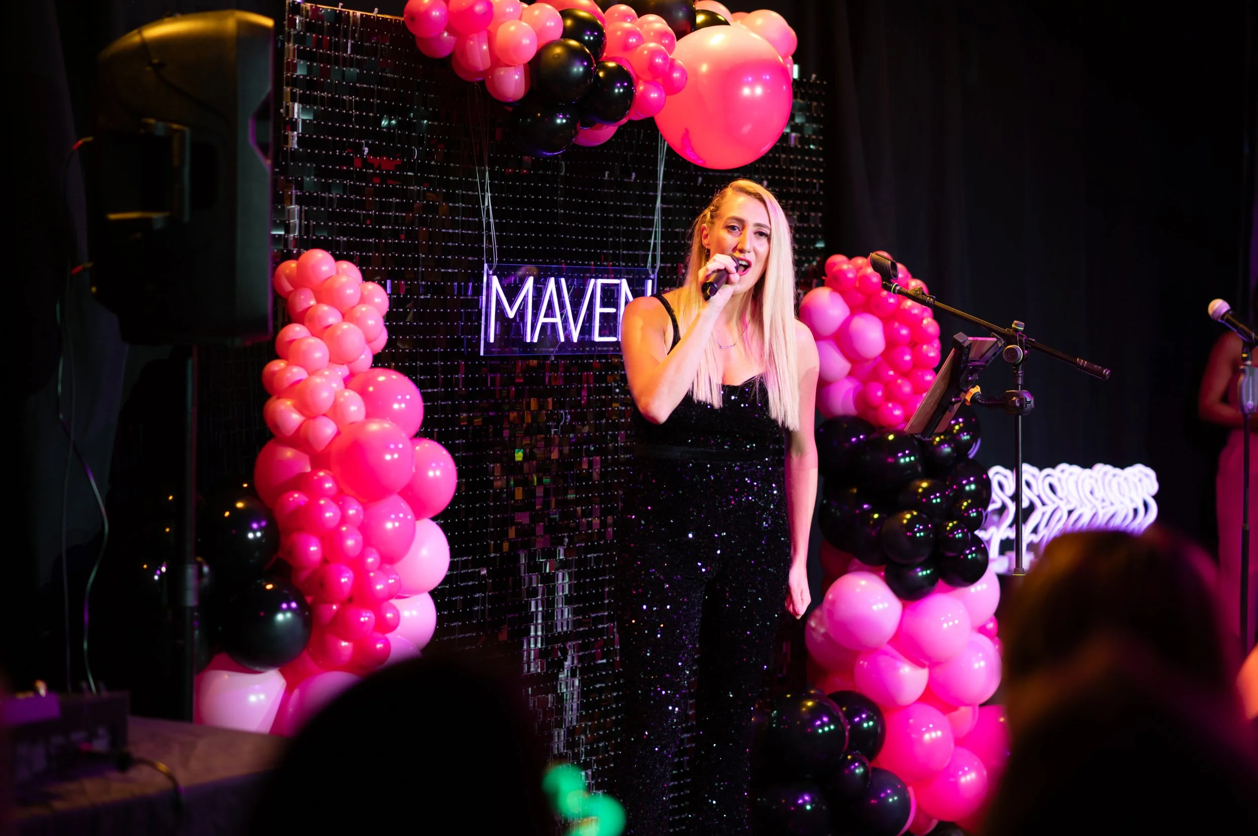A woman performing singing or speaking on a decorated stage with pink, black, and white balloons, neon sign behind her, and a black curtain backdrop.