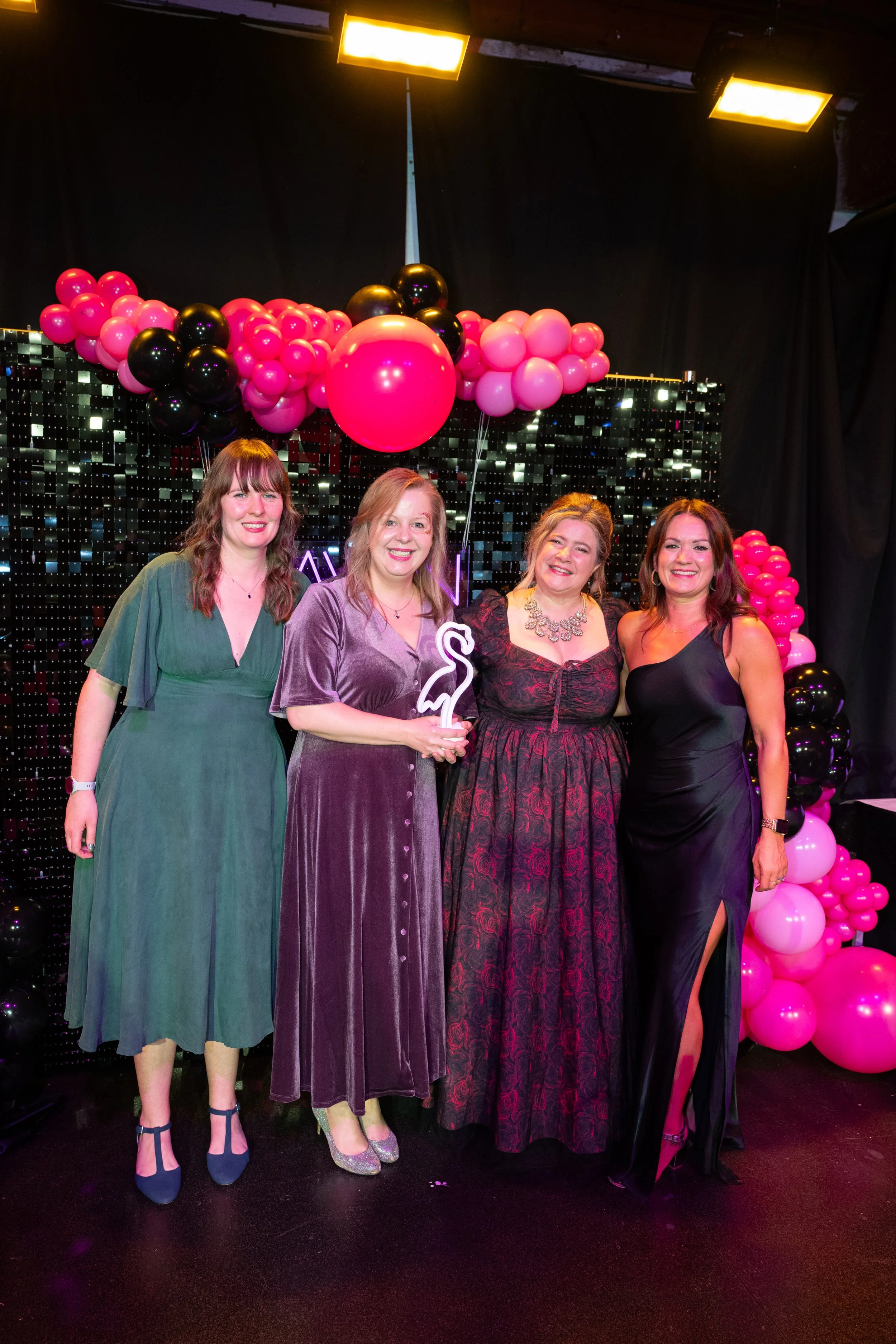 Four women standing together at a celebration event, with pink and black balloons and a black and silver backdrop behind them.