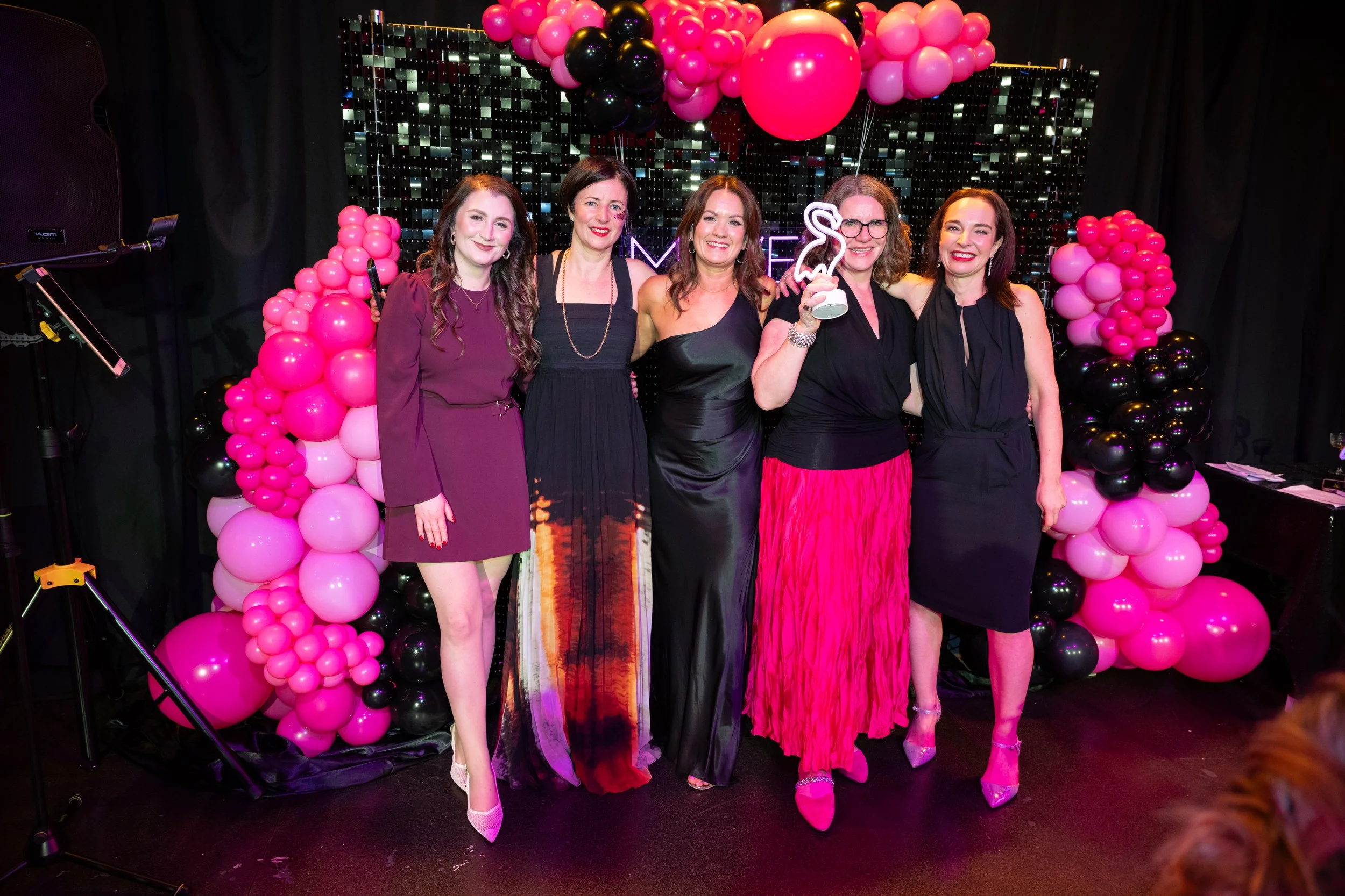Group of five women standing together at a celebration, surrounded by pink and black balloon arrangements, with a decorated backdrop and a sign that says 'M.' one woman holds a flamingo-shaped trophy. They are dressed in formal attire and smiling.