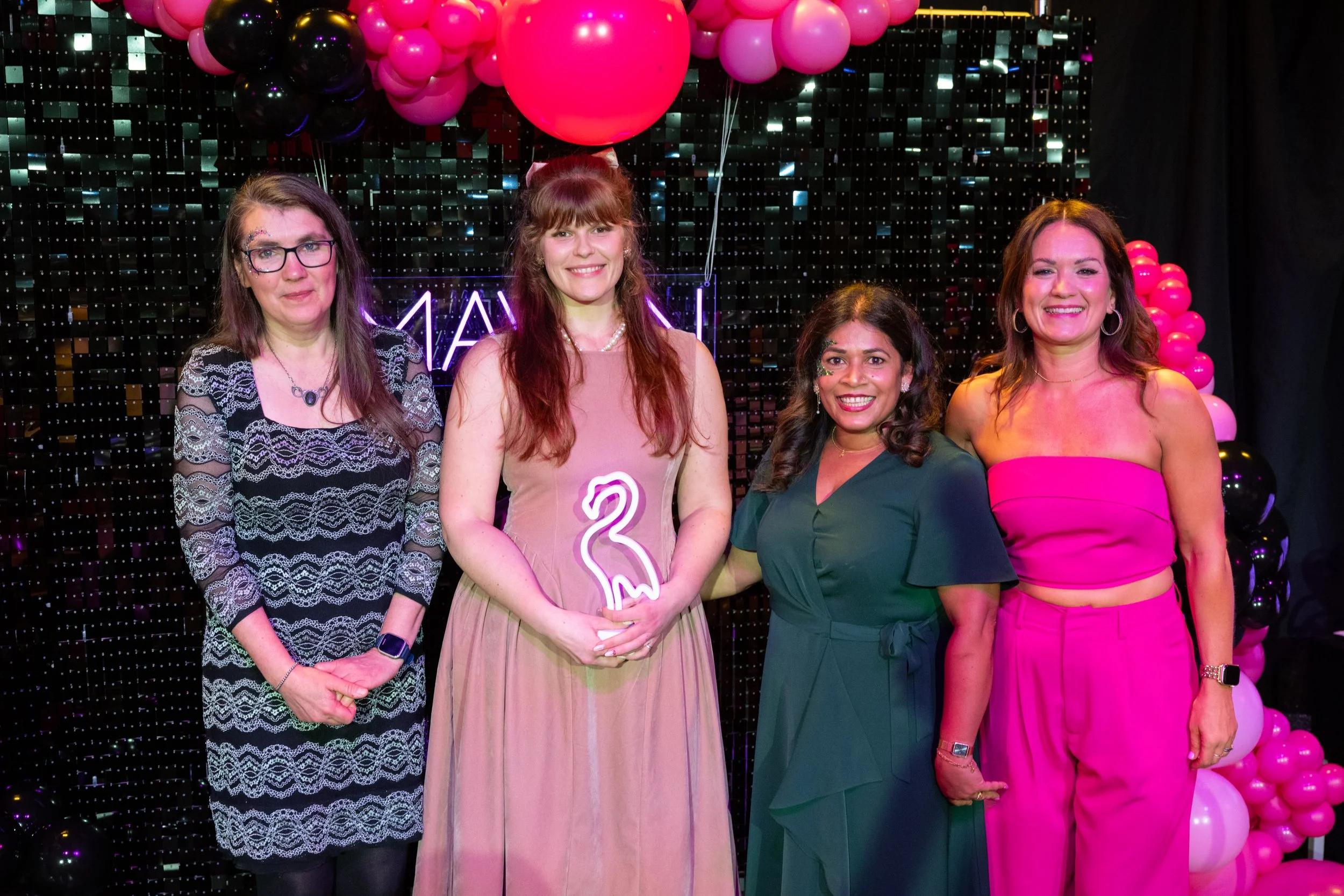 Four women standing together at a celebration event with pink and black balloons and a neon sign in the background. They are dressed in colorful outfits, smiling for the photo.