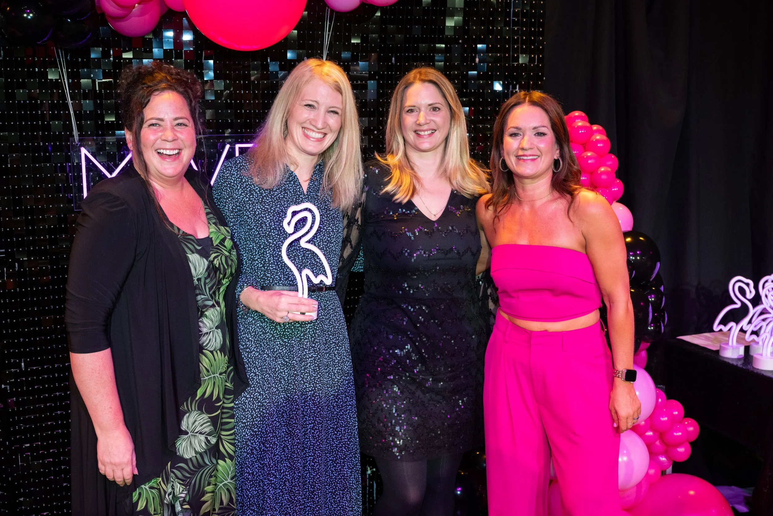 Four women standing together at a celebration or party, with black sequin backdrop, pink balloons, and pink and black balloon decorations. One woman is holding a flamingo-shaped light. They are smiling and dressed in colorful outfits.