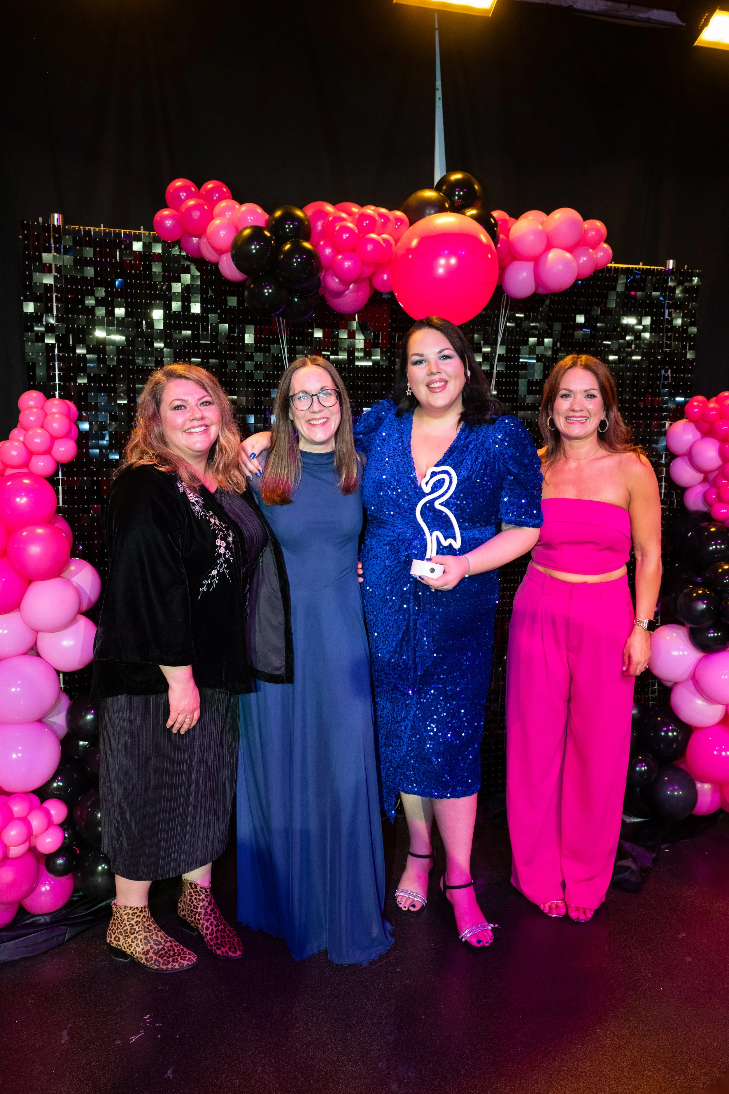 Four women standing together at a party, with a backdrop of pink and black balloons and a black shiny tiled wall. The woman second from the right holds a white candle shaped like the number 3, and all women are dressed in colorful outfits.