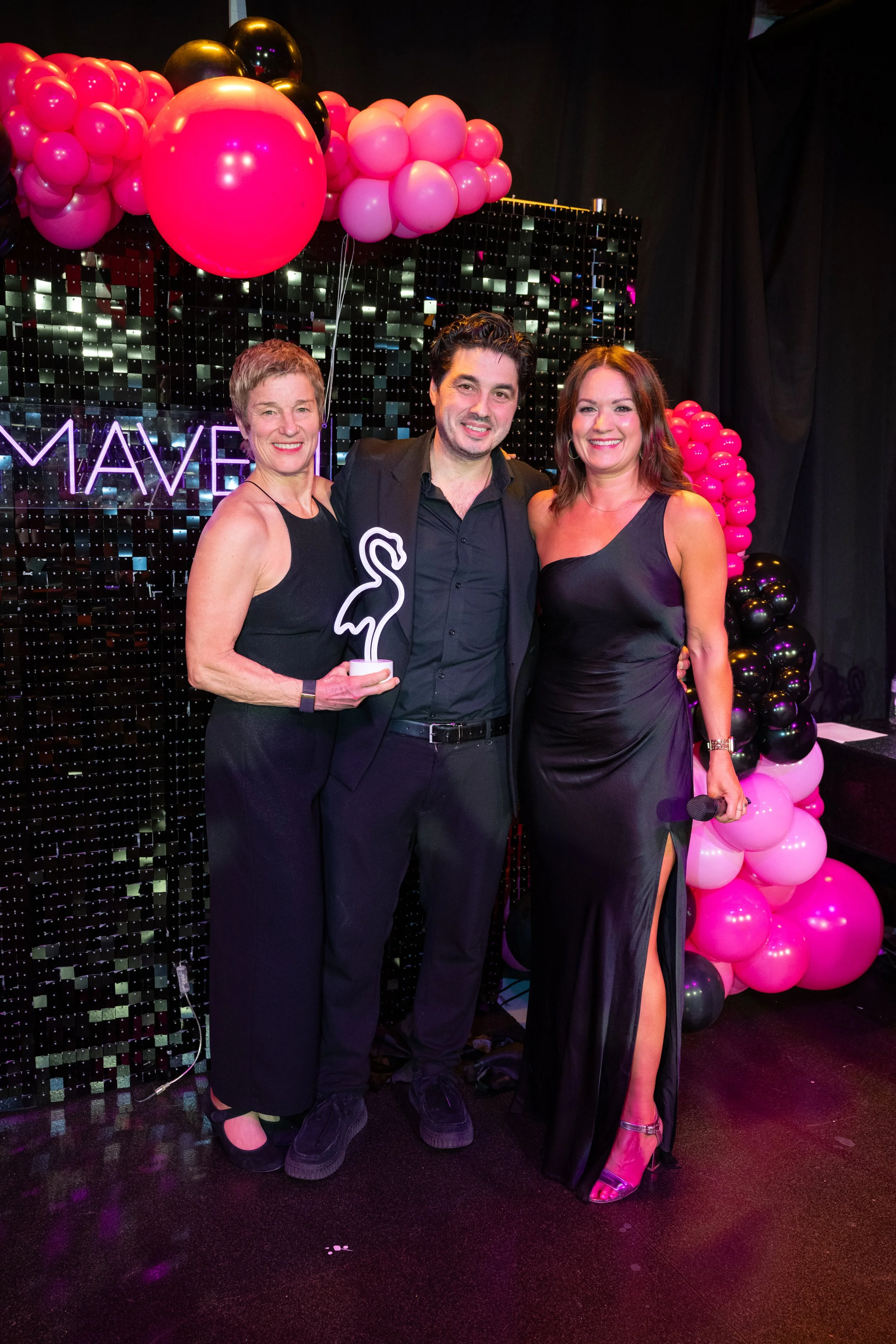 Three people dressed in formal attire at party with black and pink balloon decorations and a neon sign in the background.
