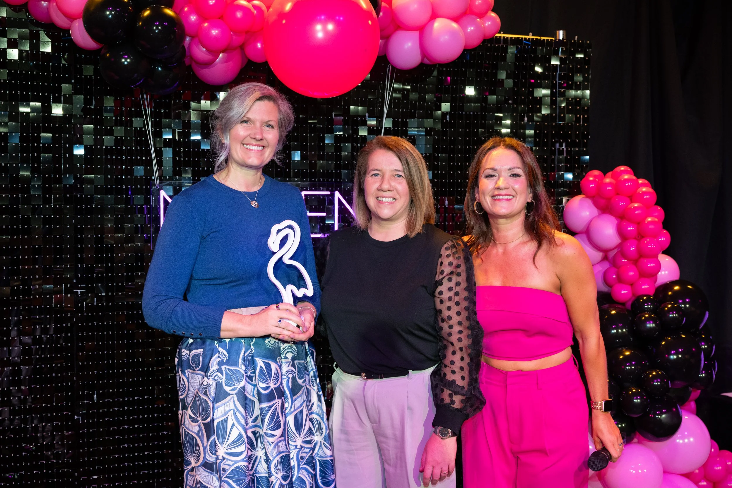 Three women standing together at a celebration with pink and black balloons and a black sparkly background, smiling at the camera.