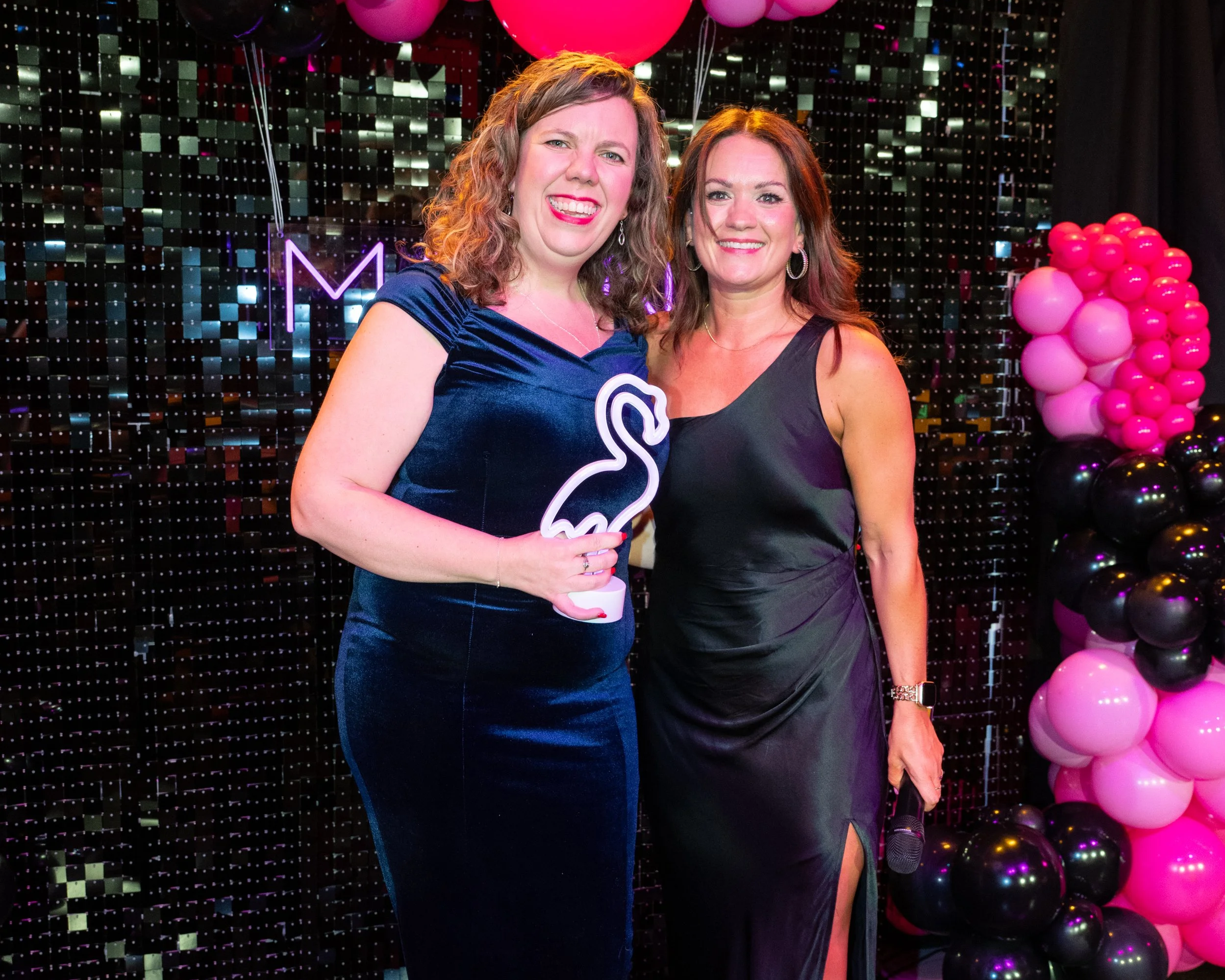 Two women in evening dresses smiling at a celebration with pink balloons and a black and pink backdrop.