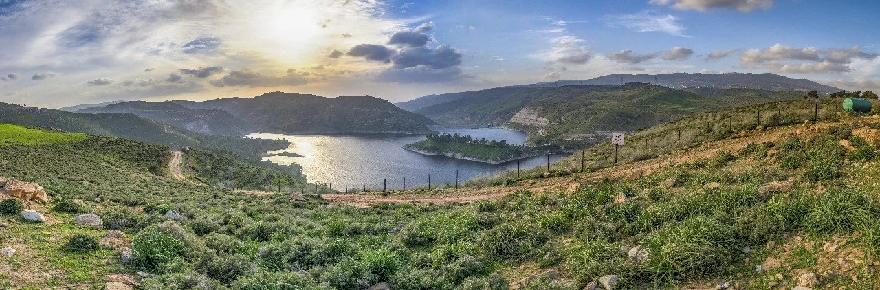 Panoramic view of a large body of water surrounded by green hills and mountains, under a partly cloudy sky at sunset.