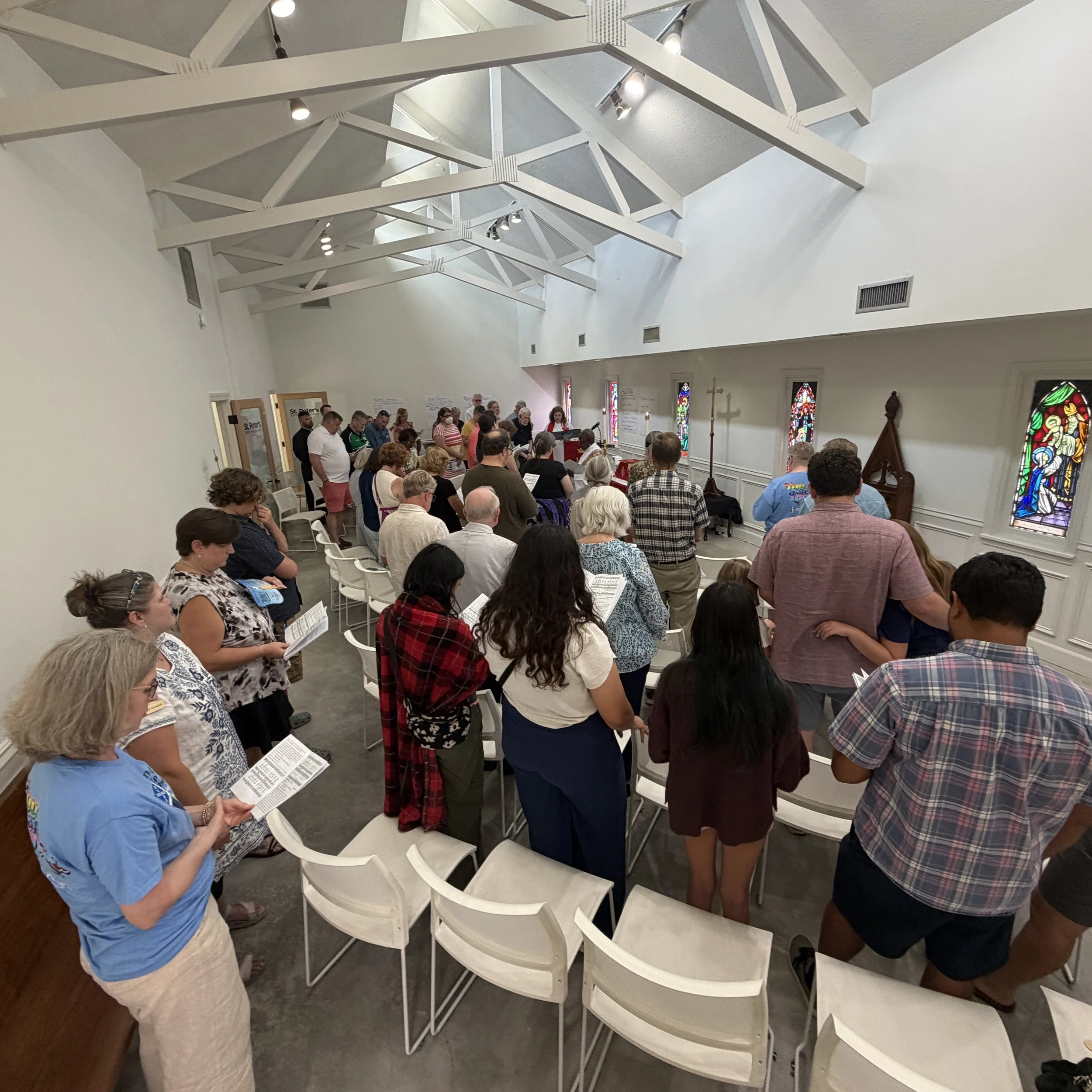 People gather in a church interior for a service or event, some reading materials, with stained glass windows and religious statues visible.