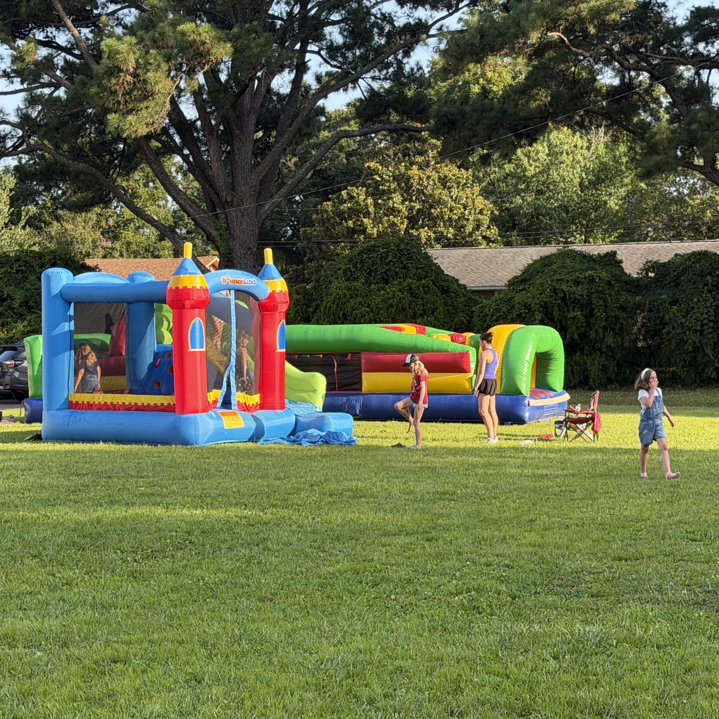 Colorful inflatable bounce house on a grassy lawn with children playing, large leafy trees and a house with a shingled roof in the background.