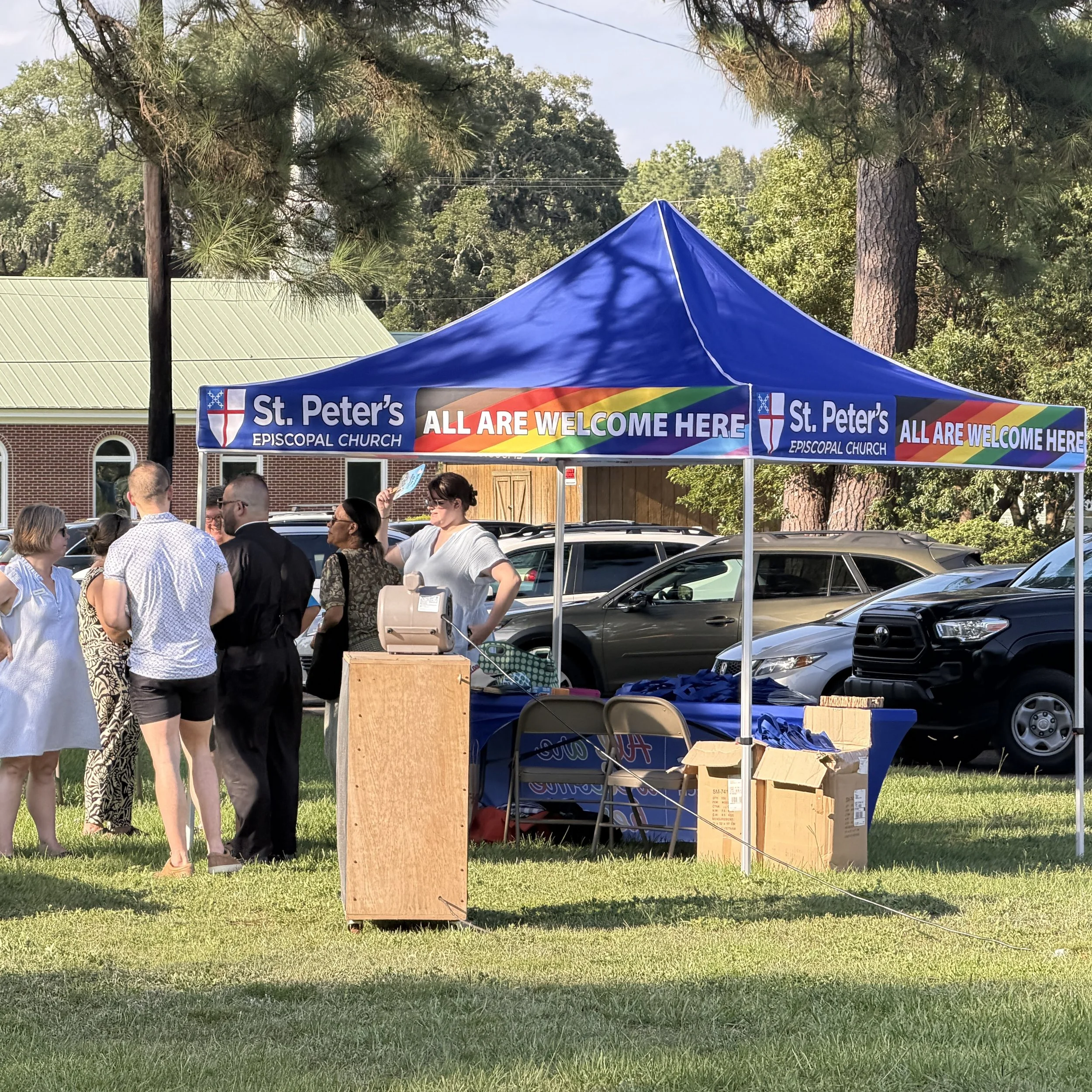 A tent with a rainbow-colored banner reading 'All Are Welcome Here' stands in a grassy outdoor area, surrounded by people and parked cars, with trees and a brick building in the background.